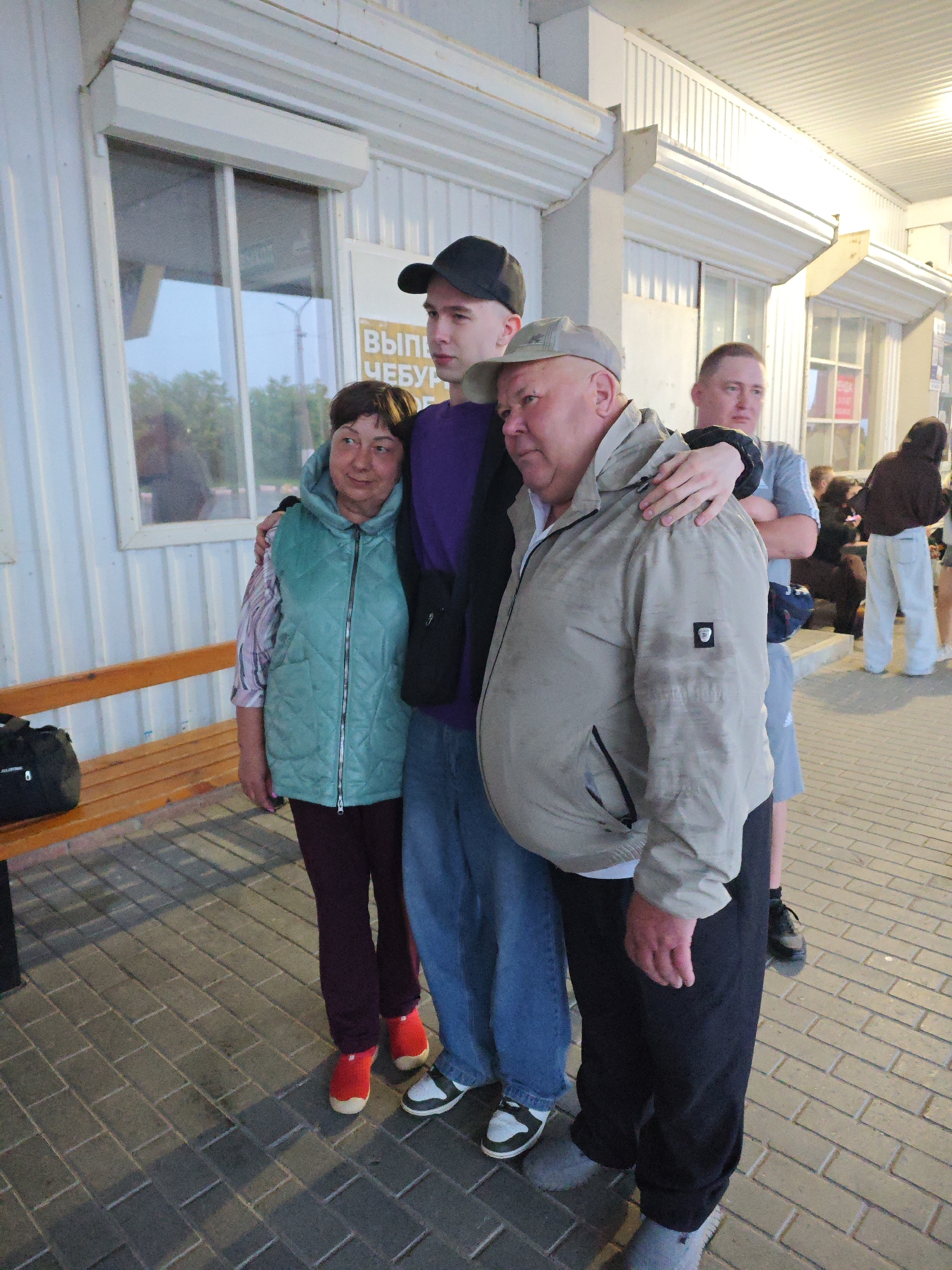Four people are standing outside a building with a white corrugated metal facade. A young man wearing a black baseball cap and a purple shirt is in the center, with his left arm around an older man in a light brown jacket and a grey cap. To the left of the young man is a woman wearing a teal quilted vest over a striped shirt and dark pants. Behind the older man, another man in a grey t-shirt is visible, with his arms crossed. In the background, other individuals are seen near shop windows. The ground is paved with grey brick. A wooden bench is to the left of the group, with a black duffel bag on it. The building appears to be a commercial structure, possibly a small shop or an open-air market. The scene suggests a casual gathering or a moment captured while passing by. The building facade suggests it might be a bus station or a similar public transit point in Chaikovsky, Russia, given the Cyrillic sign above the woman's head that reads "ВЫП... ЧЕБУ...".