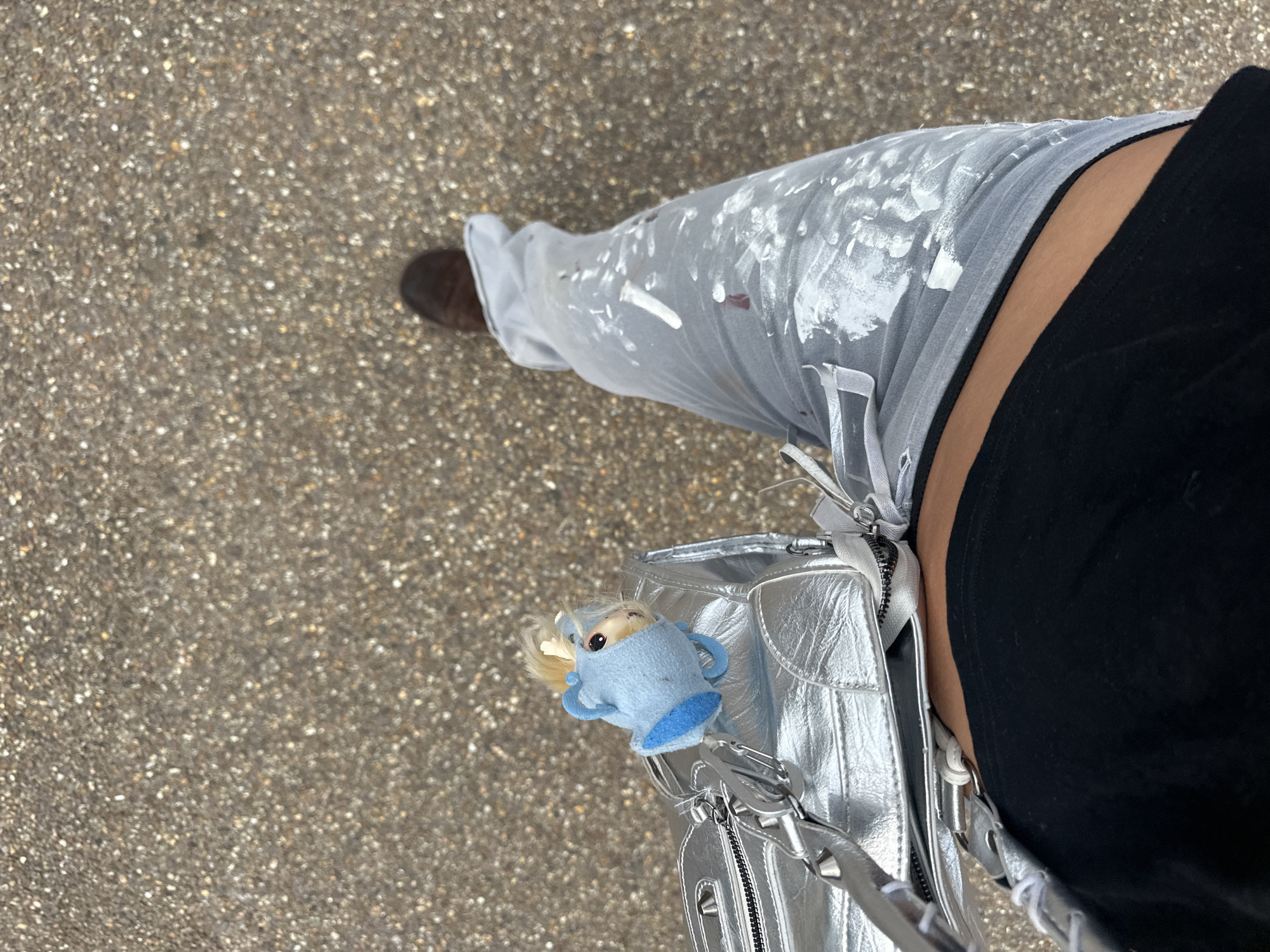 The image depicts a partial view of a person standing on a paved surface that suggests a street or sidewalk. The person is wearing light-colored pants with white paint splatters, a black top, and brown shoes. A silver handbag with a blue doll-like keychain is visible. The background is blurred and shows a paved surface consistent with a typical urban environment. The exact location in London cannot be determined from the image.