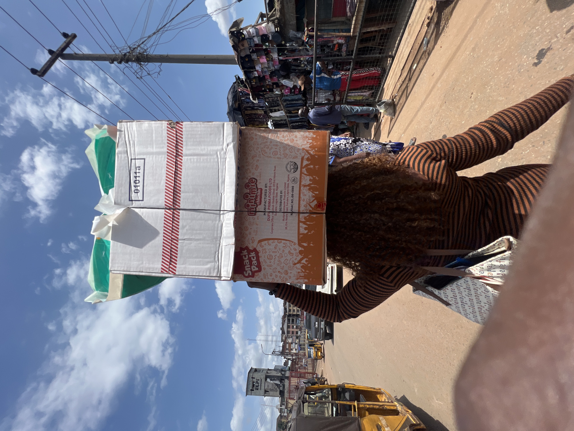 A woman in a brown and black striped shirt is balancing two boxes on her head while walking down a street in Unknown, Unknown. The top box is labeled "Snack Pack" and includes the information "DUFIL PRIMA FOODS LTD" and "minimie chinchin". The bottom box has a "01011a" label and red stripes. In the background, there's a market stall with clothes and other merchandise, a yellow taxi-like vehicle, a building with "ELPI" written on it, electrical wires, and a blue sky with scattered clouds. The street appears to be busy with pedestrian and vehicular traffic. The photographer is possibly holding a bag.