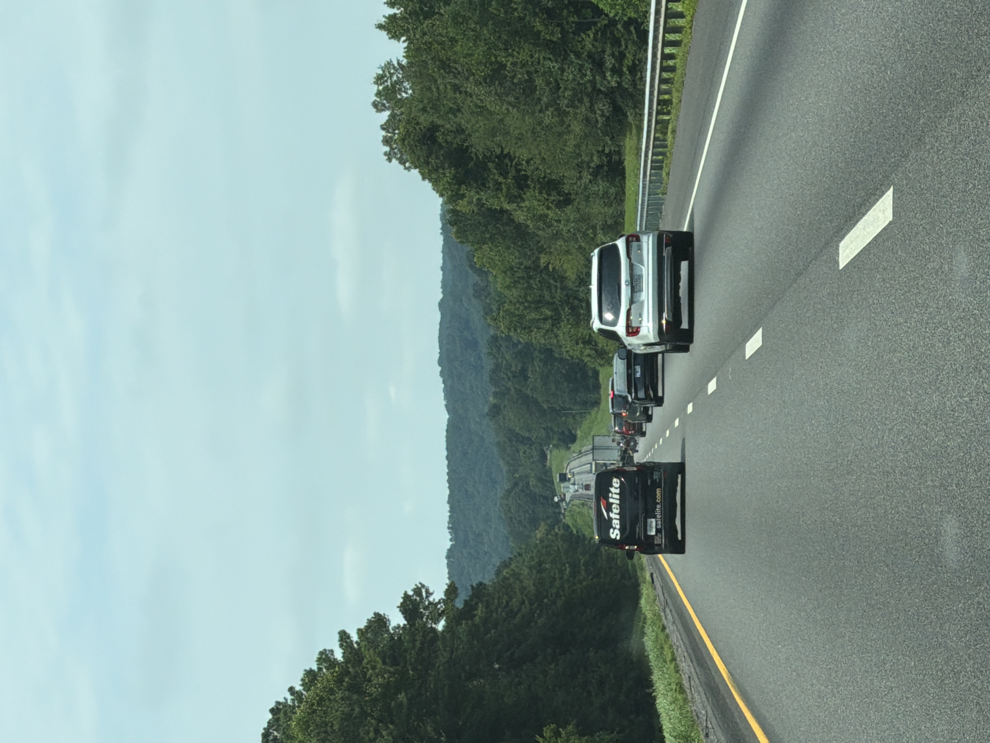 The image depicts a view from a vehicle driving on a highway. In the foreground, the asphalt road is visible with lane markings. Several vehicles are on the highway, including a black van with "Safelite" branding on its rear and a white SUV. The highway is bordered by trees and lush green foliage, with rolling hills in the background under a cloudy sky. The scene appears to be a typical highway commute in a rural or suburban area, possibly in or near McDonald, United States, given the context provided, though the exact location cannot be definitively determined from the image alone.