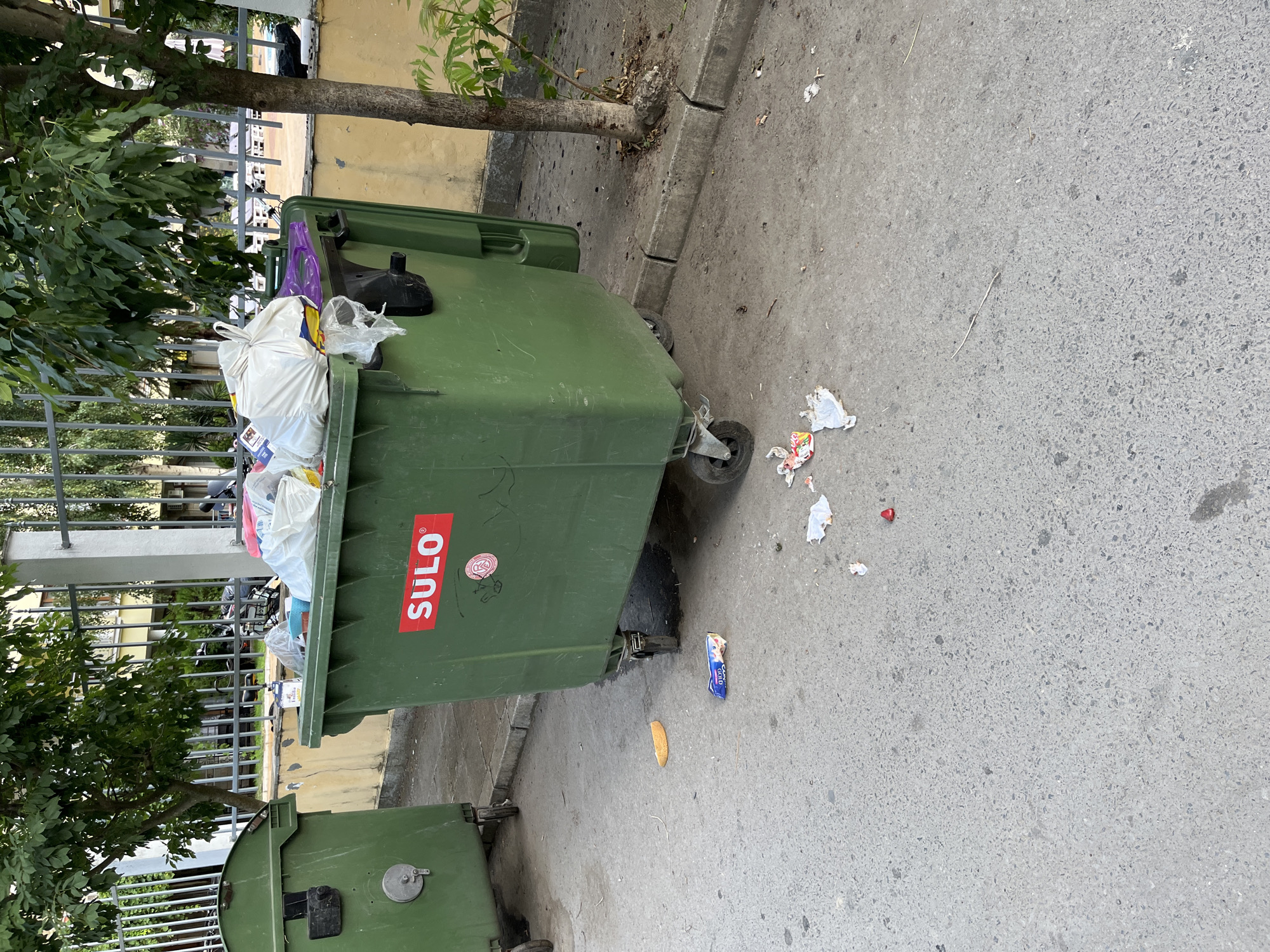 The image depicts an outdoor scene in Nesebar, Bulgaria, featuring two large green dumpsters. The dumpster in the foreground is overflowing with trash, including white plastic bags, and has a red "SULO" logo on its side. Debris is scattered on the asphalt ground around the dumpsters. A piece of what appears to be a bread crust or pastry is visible on the ground to the left of the foreground dumpster, along with a crumpled wrapper. Behind the dumpsters, a metal fence and a building with yellow siding are visible. Trees with lush green foliage are present, casting shadows on the scene. The overall impression is one of a public area with waste management facilities.