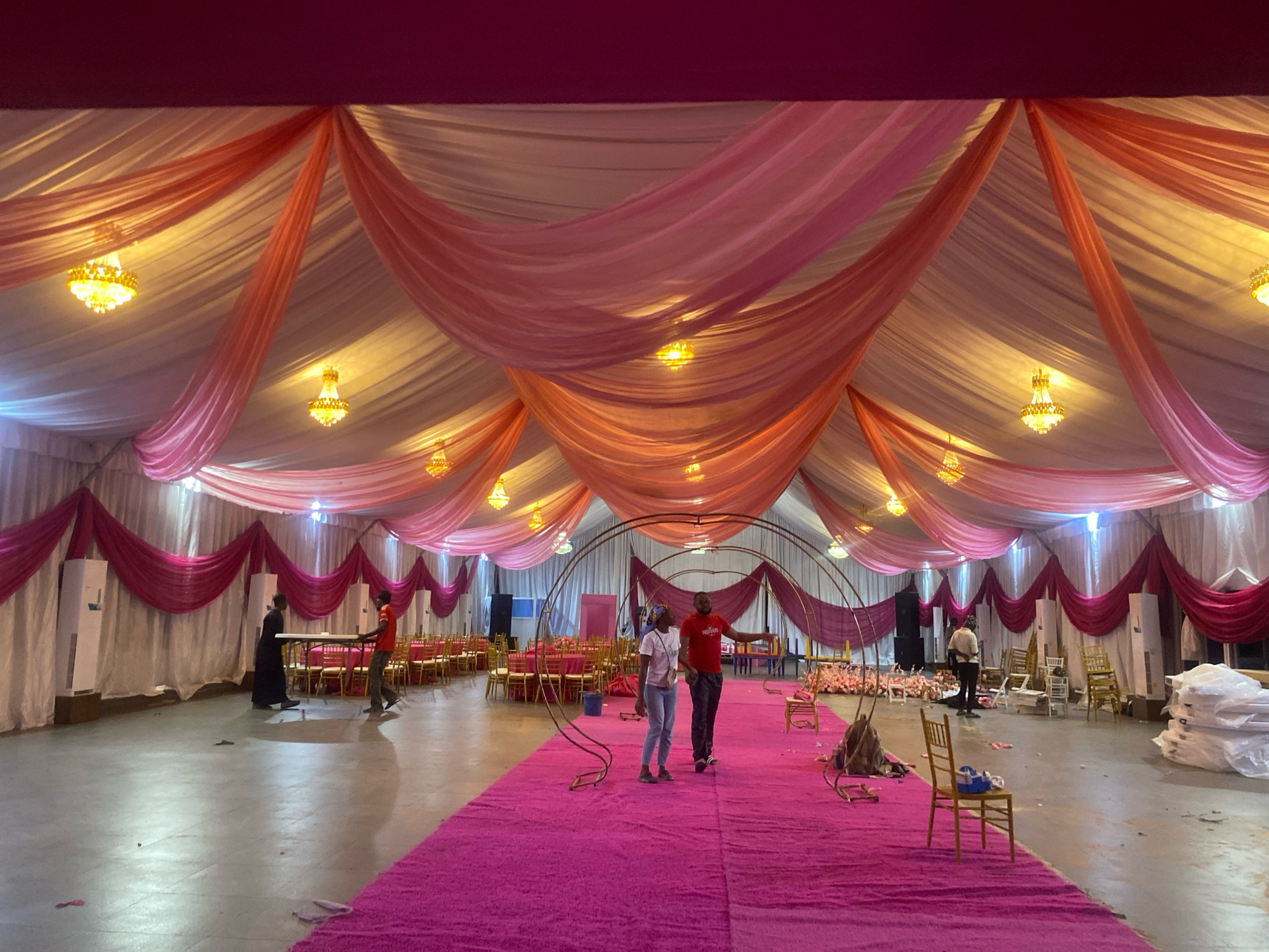 The image depicts the interior of a large tent or event hall in Barden Yana, Nigeria, currently undergoing setup for an event. The ceiling is draped with layers of white and pink fabric, creating a voluminous and decorative effect. Several ornate chandeliers hang from the ceiling, emitting a warm light. A long, bright pink carpet runs down the center of the hall.
In the foreground, two individuals, a man and a woman, stand on the pink carpet, appearing to be observing or directing the setup. The woman wears a white t-shirt and blue jeans, while the man is in a red t-shirt and dark pants. They are positioned near a large, circular metal archway, one of several that are part of the event's decor.
Further into the hall, several other people are visible engaged in various setup tasks. One person in a black outfit walks near a table, and another individual in a red shirt is seen near a setup area with chairs. The hall is furnished with rows of gold-colored chairs and tables, some of which are still being arranged or set up. In the background, stacks of white materials, possibly linens or more decor, are visible, along with speakers and other event equipment. The overall impression is one of active preparation for a celebratory occasion.