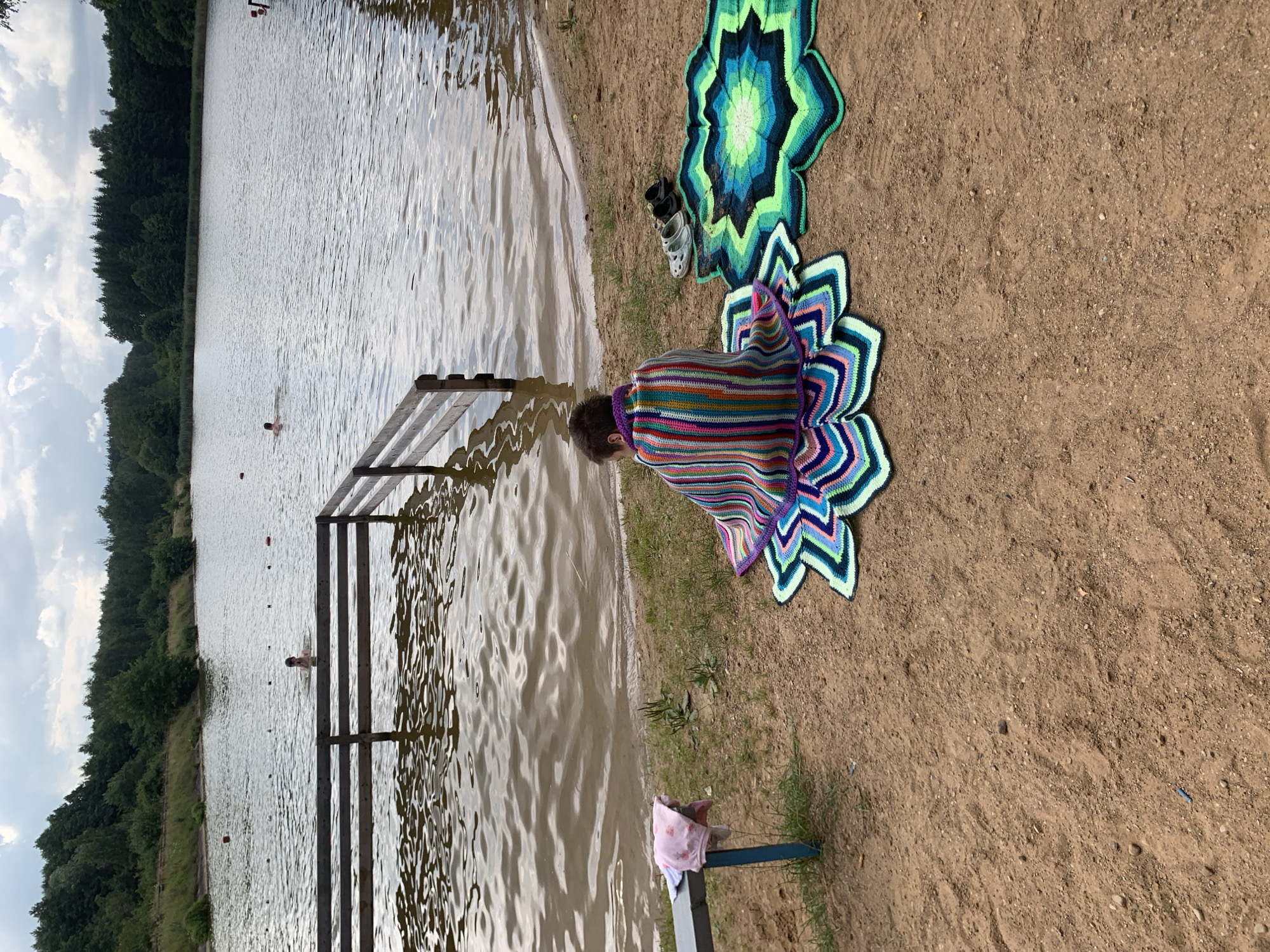 A young person, facing away from the camera and wrapped in a colorful striped blanket, sits on a lotus-shaped crocheted mat on a sandy shore. Beside them, another crocheted mat in shades of green and blue is laid out, along with a pair of sandals. A blue bench with a pink cloth is to the left. In the background, a lake is visible with a wooden barrier separating the shore from the water. Two individuals are swimming in the lake, and a tree-lined shore is seen in the distance. The scene is set in a natural outdoor environment, likely a beach or lakefront area within the Грозаўскі сельскі Савет region of Belarus.