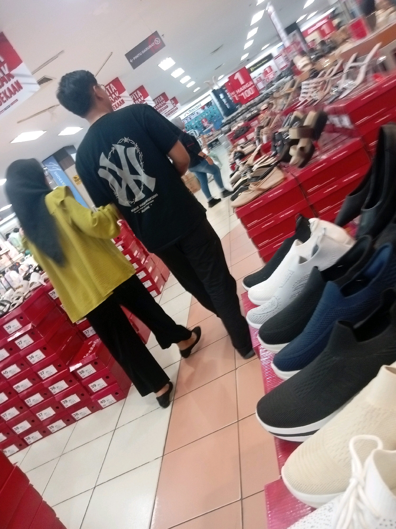 The image depicts an indoor scene, likely a retail store, with rows of shoe boxes and display shelves filled with footwear. In the foreground, a man wearing a black t-shirt with a graphic print and black pants is facing away from the camera. To his left, a woman wearing a yellow long-sleeved top and black pants, with a black hijab, is also facing away. They appear to be browsing or walking through the store. The environment shows bright overhead lighting and tiled flooring. Red shoe boxes are stacked prominently, some with visible labels indicating shoe sizes. In the background, more shoes are displayed on shelves, and other shoppers are visible. The overall atmosphere suggests a typical shopping experience in a large retail establishment. This scene is likely from a shopping mall or department store in Batam, Indonesia, given the context of the image.