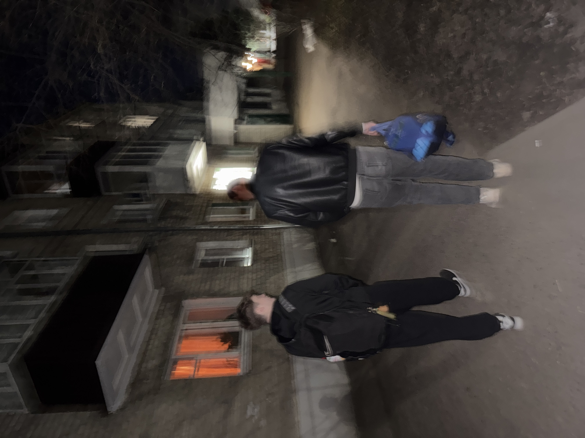 The image captures a blurry, low-light outdoor scene at night in Kazan, Russia, depicting two individuals walking along a paved urban path. The image appears rotated 90 degrees counter-clockwise from its original orientation.
The person on the left (when upright) is a male, seen from behind, wearing a black leather jacket, grey jeans, and white sneakers. He holds a blue backpack in his left hand. The second person, slightly behind and to the right, also appears male, dressed in a dark jacket or hoodie, black pants, and light-colored sneakers. A small bright yellow or orange object, possibly keys, hangs from his hip. Both figures are rendered with significant motion blur, suggesting they are in movement or the photo was taken with a slow shutter speed in dim light.
In the background, to the left of the path, is the blurry facade of an apartment or commercial building made of light-colored brick. Numerous windows are visible, some glowing with warm orange and bright white interior lights. Above the building, bare tree branches are silhouetted against the dark night sky. The ground under the individuals is a dark, uniform paved surface. No discernible text is visible anywhere in the image. The overall impression is a candid, gritty street photograph taken during nighttime hours.