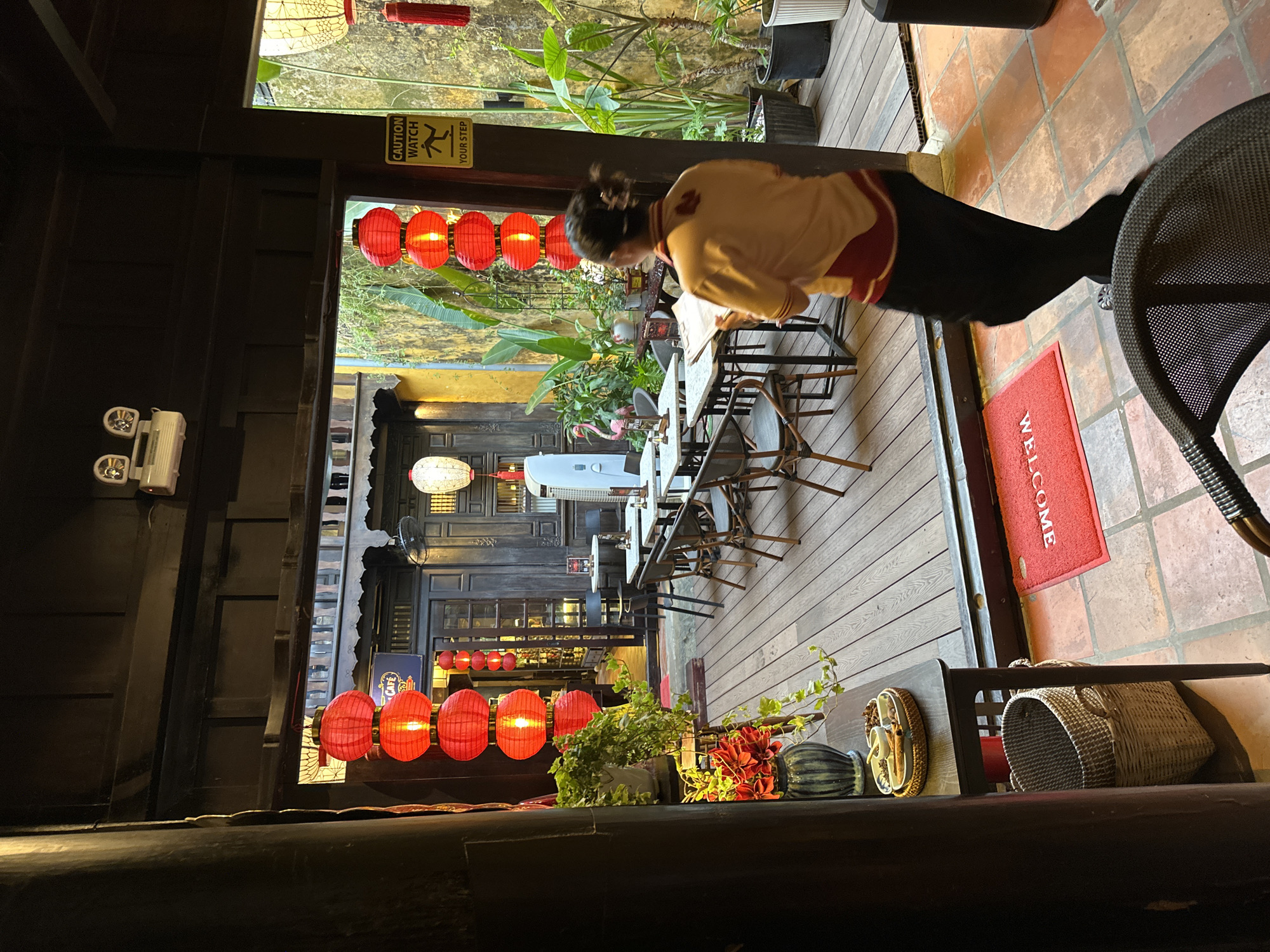 The image depicts an outdoor seating area of a cafe in Hội An Ward, Vietnam, at night. Red lanterns are suspended from the building's eaves, casting a warm glow on the scene. A woman wearing a yellow and red uniform is walking away from the camera, holding a menu or papers. She is in the foreground, on the right side of the image. The seating area features wooden decking and is furnished with tables and chairs. In the background, a traditional building with dark wooden doors and window frames is visible. Lush green plants are arranged throughout the outdoor space, and a "CAUTION WATCH YOUR STEP" sign is posted on a pillar. A red welcome mat is placed on the tiled floor near an entrance. The atmosphere is dimly lit, suggesting the establishment is open for evening service.