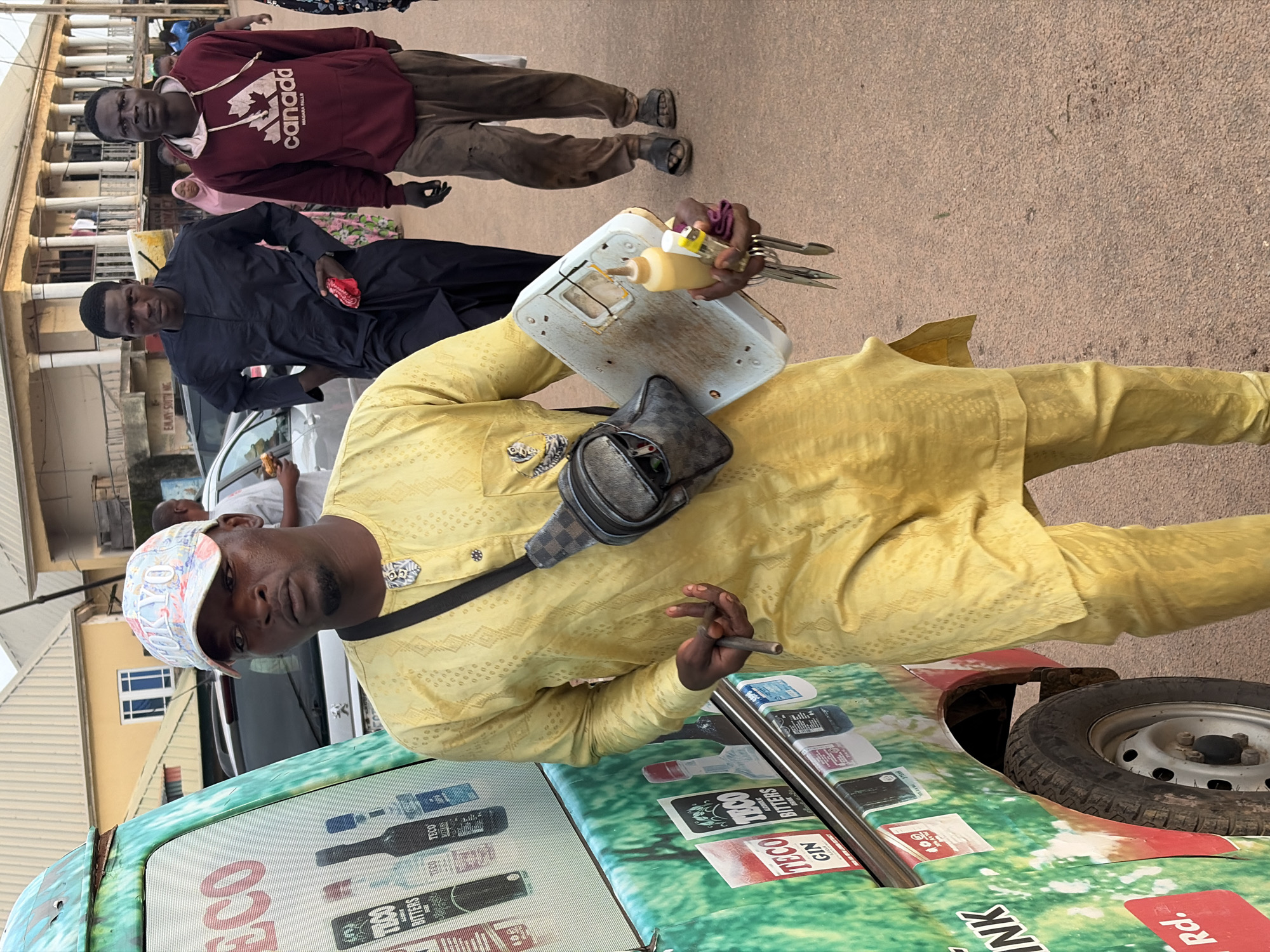 In Gura Top, Nigeria, a man in a yellow traditional outfit and a "Tokyo" baseball cap stands in front of a green vehicle with colorful branding. He holds a small scale in his right hand, which appears to be used for weighing goods. In his left hand, he holds what looks like a cigar. Behind him, two men walk away from the camera; one in a black kaftan and the other in a maroon hoodie with "Canada" printed on it. The background features buildings with archways and columns, characteristic of the region's architecture, and suggests a public street or marketplace. The lighting indicates it's daytime, possibly late afternoon, given the long shadows.
