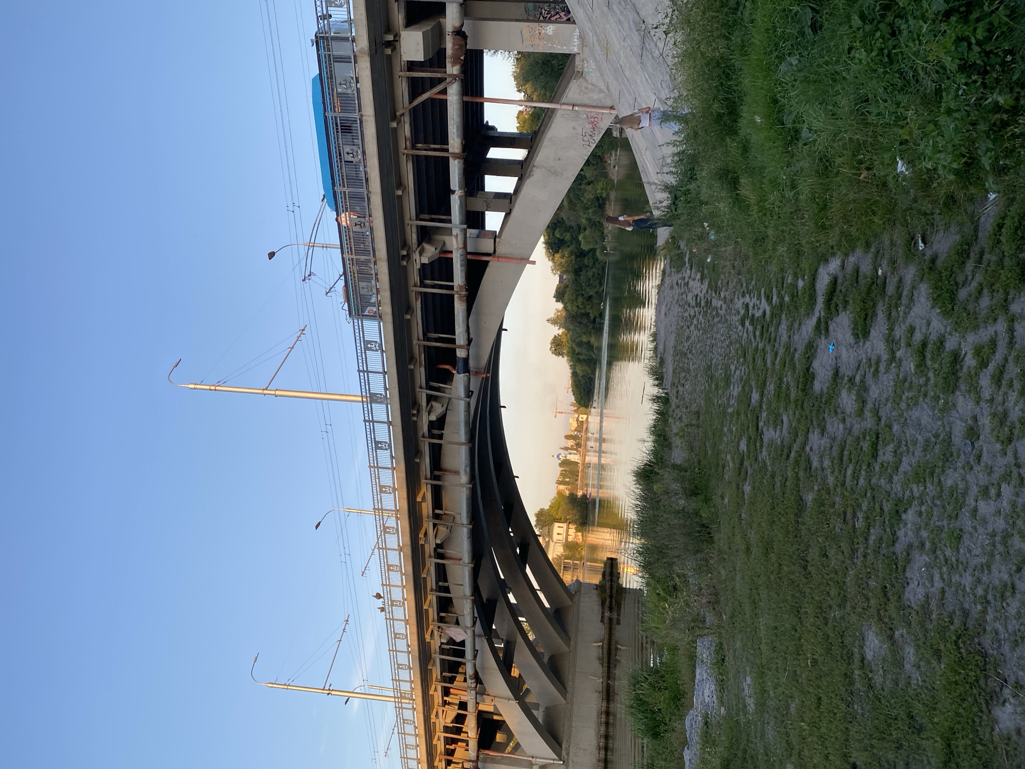 The image captures a view of the trolleybus line crossing over the Southern Bug River in Vinnytsia, Ukraine. The sun is setting, casting a warm glow on the scene. In the foreground, there is a patch of grass and a sandy area leading down to the riverbank. Two young women are standing on the bank, looking towards the river. The trolleybus line, a prominent structure, spans across the river, with a blue trolleybus visible on the tracks. The river itself is calm, reflecting the sky and the surrounding greenery. In the background, buildings and trees line the opposite bank of the river. The overall atmosphere is serene, with a touch of urban life visible through the presence of the trolleybus and the infrastructure of the bridge.