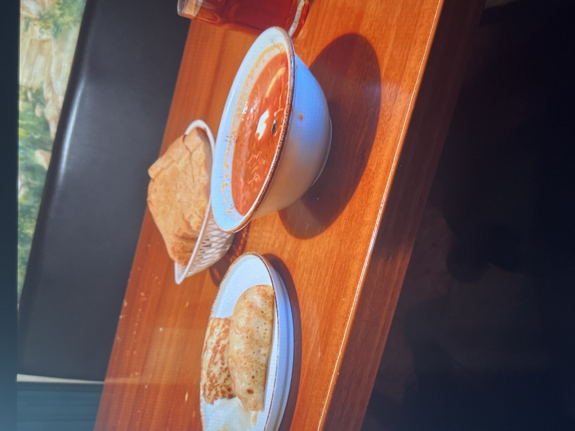 The image shows a close-up, eye-level shot of a wooden table set with food. On the left, a white plate holds two rolled crepes. In the center, a white basket contains several pieces of toasted bread. To the right, a white bowl is filled with red soup topped with a dollop of white cream. A glass of dark red liquid, likely juice or tea, stands to the far right. The background features a dark upholstered bench and a mural depicting a natural landscape. The scene appears to be set in a restaurant in Krasnoyarsk, Russia, possibly at a cafe or dining establishment.