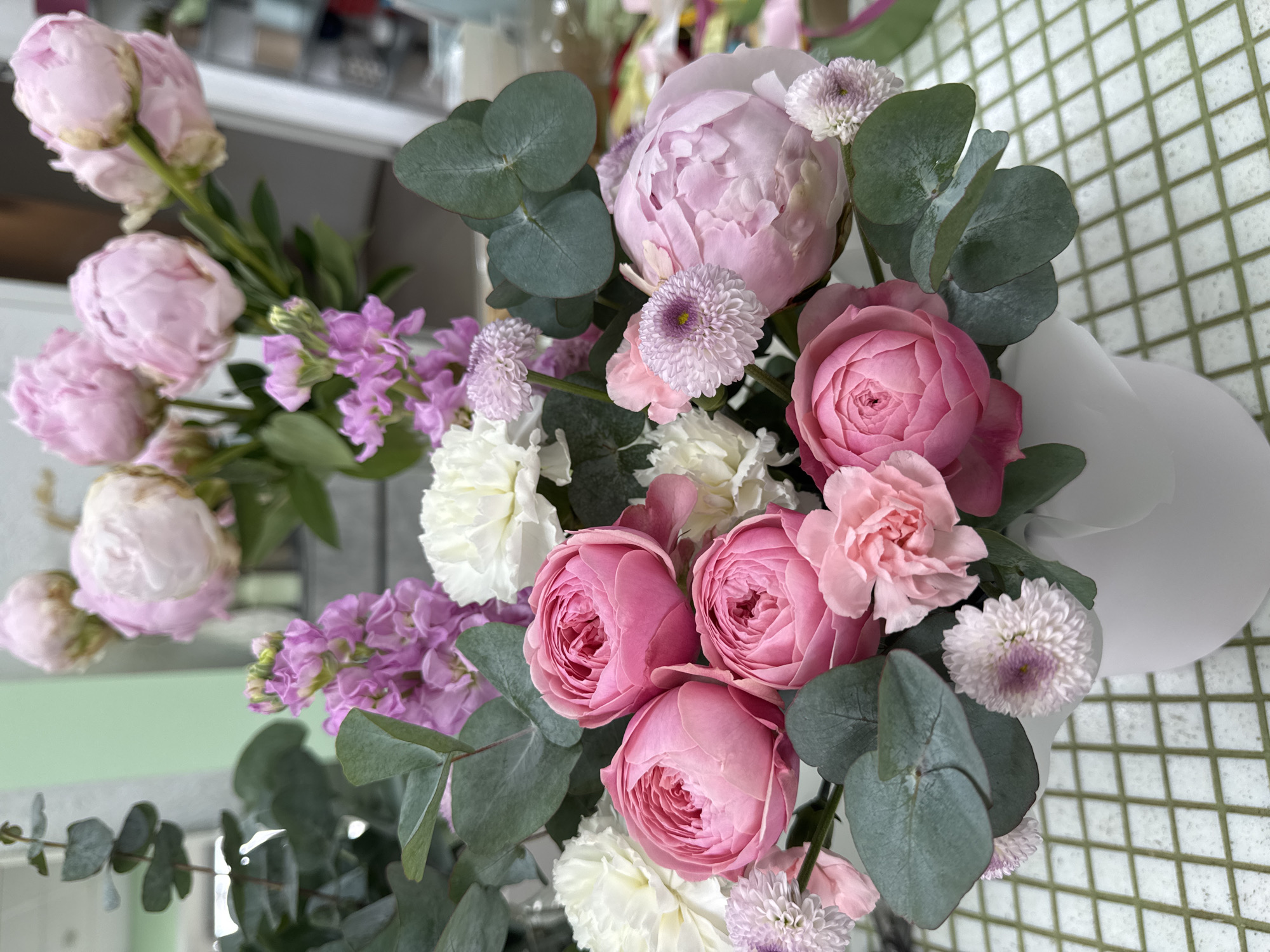 The image shows a bouquet of pink and white flowers, likely peonies, roses, carnations, and small chrysanthemums. There is also some purple stock and eucalyptus leaves in the arrangement. The bouquet is placed in a white container on a surface that appears to be tiled. The location, Izhevsk, Russia, cannot be determined from the image alone.