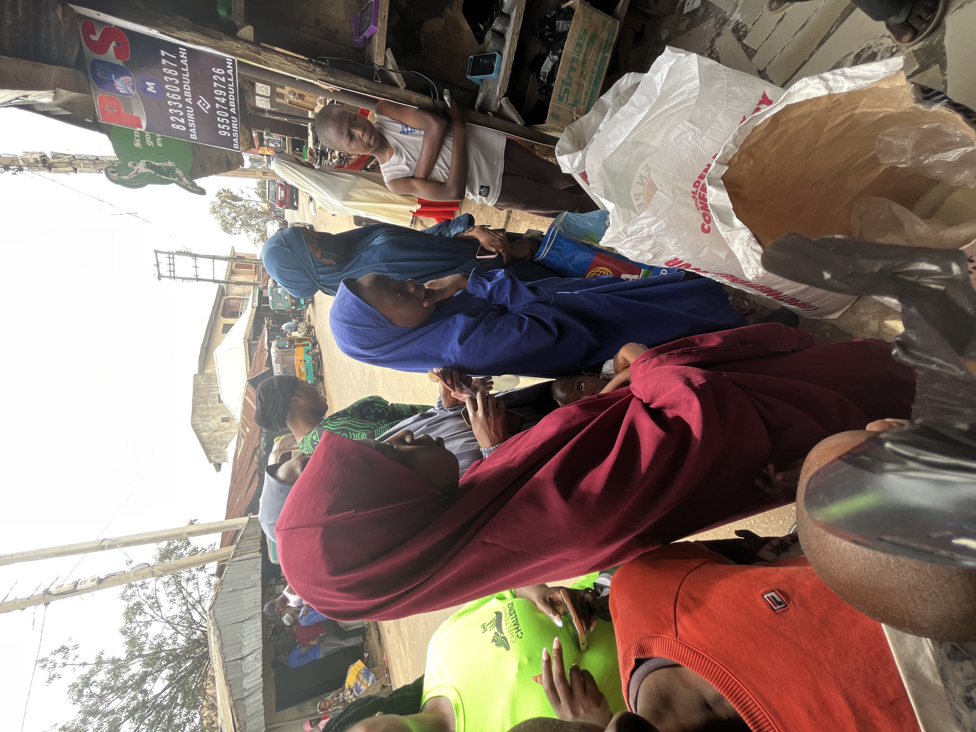 The image depicts a scene in what appears to be a street in Jos, Nigeria. A group of people, several women wearing hijabs of various colors (red and blue), are gathered. There are other individuals including a young boy in a white shirt leaning against a structure. A large white bag is also visible. In the background, a sign displays the word "POS" with phone numbers (8233603877 and 9550749726) and names "BASIRU ABDULLAHI." The environment seems to be a bustling street with visible buildings, power lines, and vehicles in the distance. A neon green shirt with the words "CHELTENHAV CHALLENGE" is also visible.