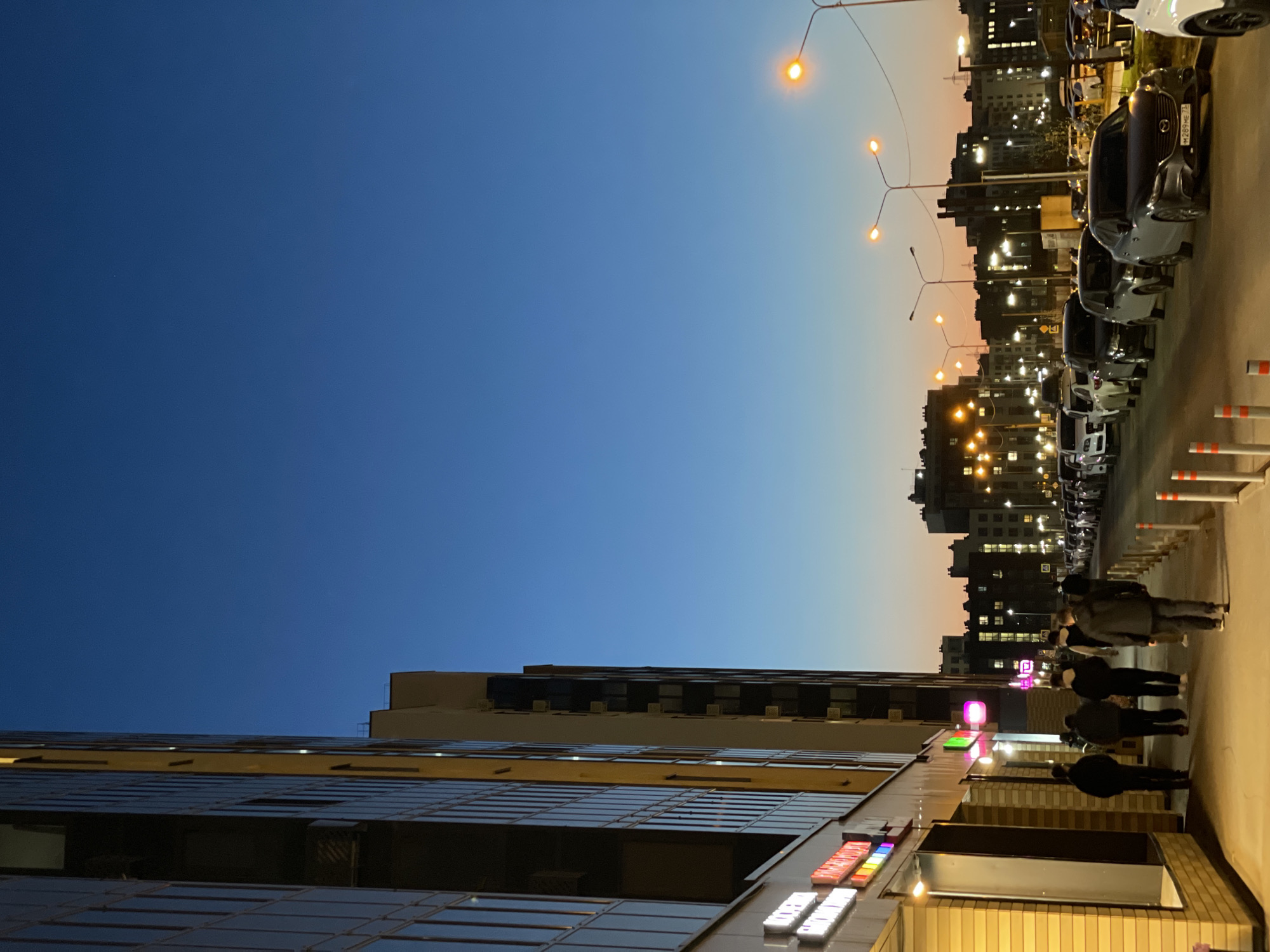 The image depicts a twilight scene in Kazan, Russia. In the foreground, the bottom of a modern building with glass panels and a few lit signs is visible. A group of people is walking on the sidewalk next to the building. Cars are parked along the street, lit by streetlights. In the background, taller buildings with lighted windows can be seen against the sky. The sky transitions from a deep blue to a lighter orange gradient near the horizon.