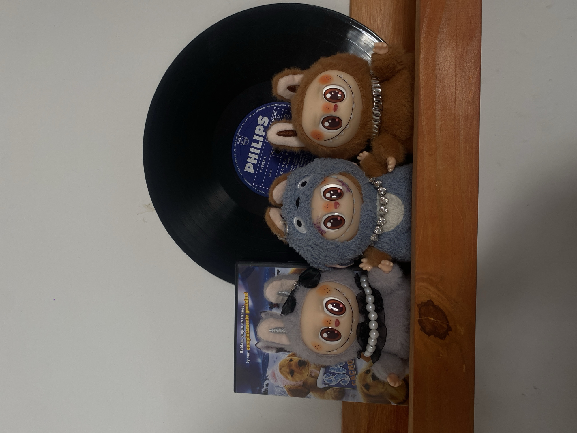 A close-up, studio shot features three plush toys stacked on top of each other, a vinyl record, and a DVD case, all resting on a wooden shelf. The toys, resembling characters with large, cartoonish eyes, are arranged vertically. The top toy is brown, the middle one is grey-blue, and the bottom one is grey. Each toy is adorned with a necklace. Behind the toys is a black vinyl record with a blue "PHILIPS" label visible at the center. The spine of a DVD case is visible at the bottom-left, reading "Hablan, viajan en trineos iy son completamente geniales!" along with "CACHO" and other Spanish words. The background is a plain white wall. The shelf, made of light-brown wood, has a vertical orientation.