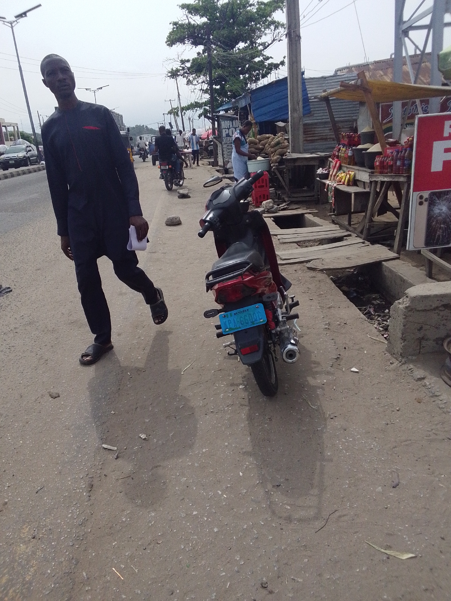 The image depicts a street scene in Lokoja, Nigeria. A man wearing a dark-colored traditional garment and sandals walks along a rough, paved road. He carries a white paper in his hand. Next to him is a parked motorcycle with a blue license plate reading "KRA-660QC" and "KOGI NIGERIA" visible. In the background, there are market stalls with vendors and various goods displayed, including drinks, food items, and what appears to be yams. People are walking and riding motorcycles in the distance. A drainage ditch partially covered with wooden planks runs along the roadside near the stalls. The sky is overcast.