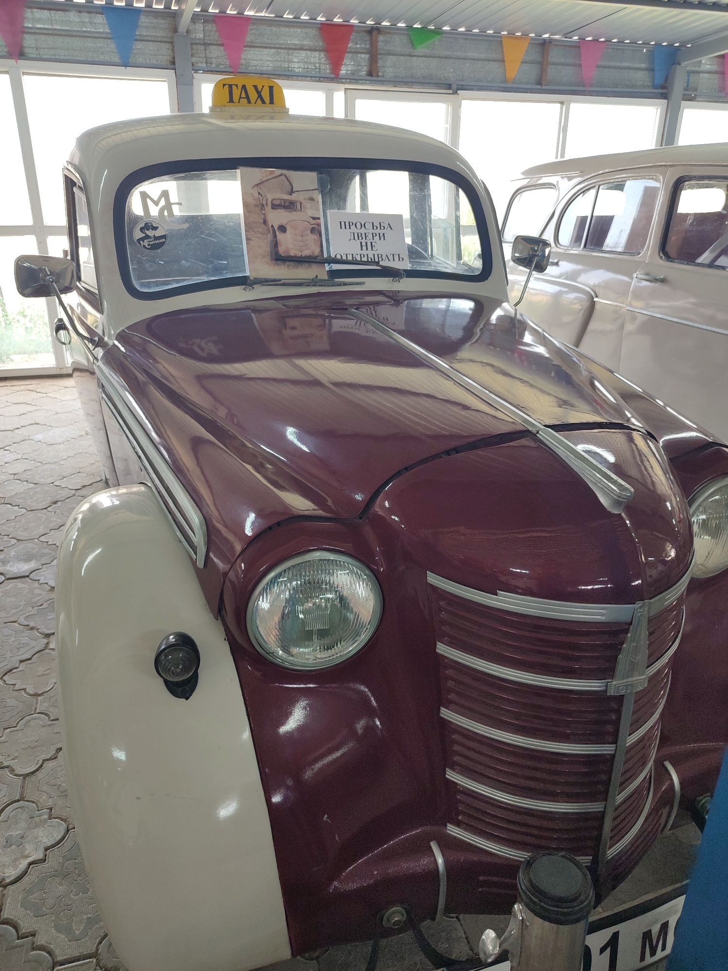 A vintage taxi, identified by a yellow "TAXI" sign on its roof, is parked indoors, possibly in a museum or exhibition space, with other classic cars visible in the background. The taxi is a two-tone vehicle, with a cream-colored top and fenders, and a deep maroon body. Its grille is distinctive, featuring horizontal chrome-colored bars. A sign on the windshield reads "ПРОСЬБА ДВЕРИ НЕ ОТКРЫВАТЬ," which translates to "PLEASE DO NOT OPEN THE DOORS." The car is parked on a patterned tiled floor. The overall setting suggests a display of vintage automobiles.