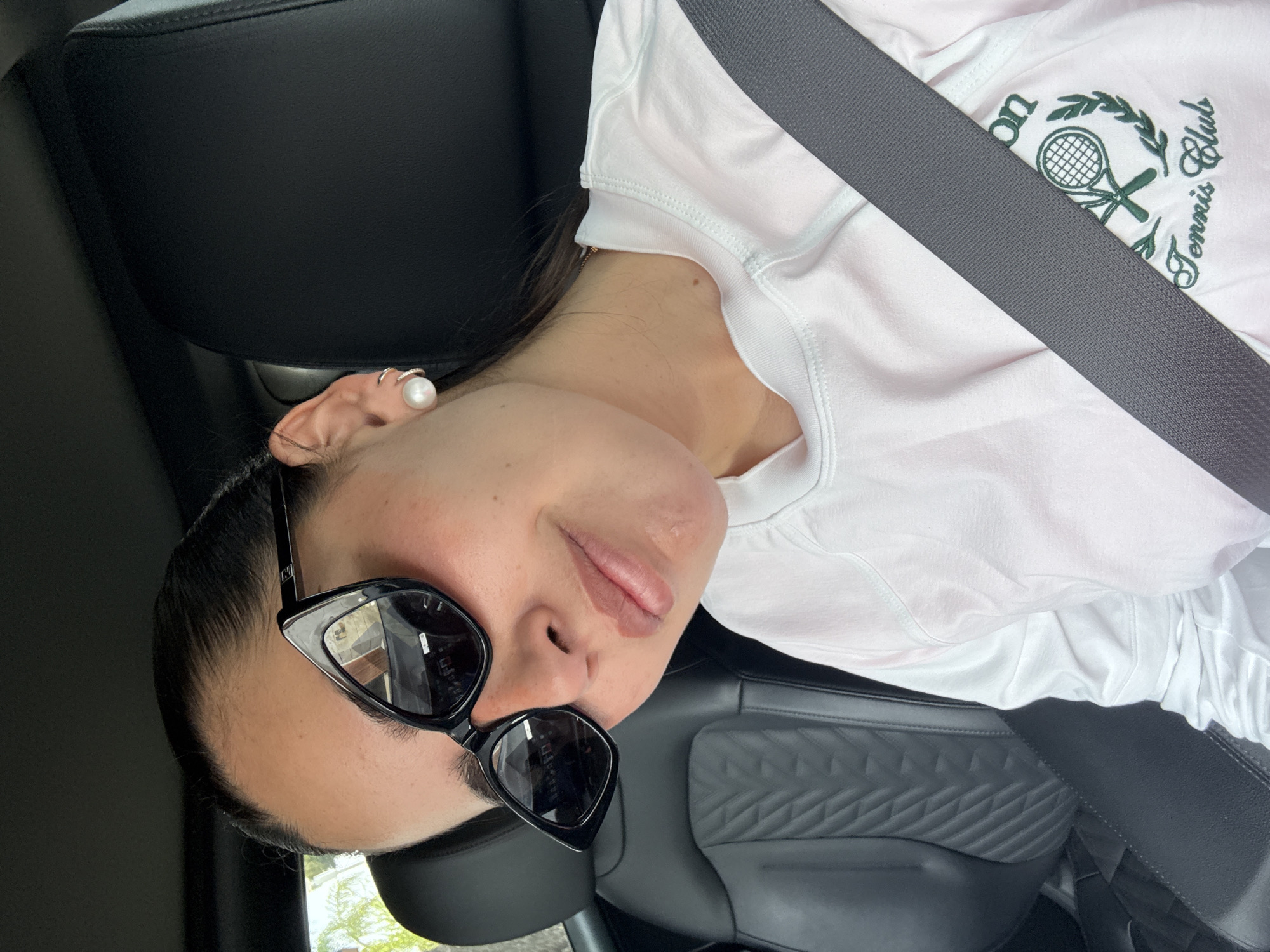 A woman wearing sunglasses and a white T-shirt with "Tennis Club" embroidered on it is sitting in a car. She has a seatbelt on and a pearl earring in her left ear. The car's interior has black leather seats with a quilted pattern. The photo was taken in San Bernardino Tlaxcalancingo, Mexico.