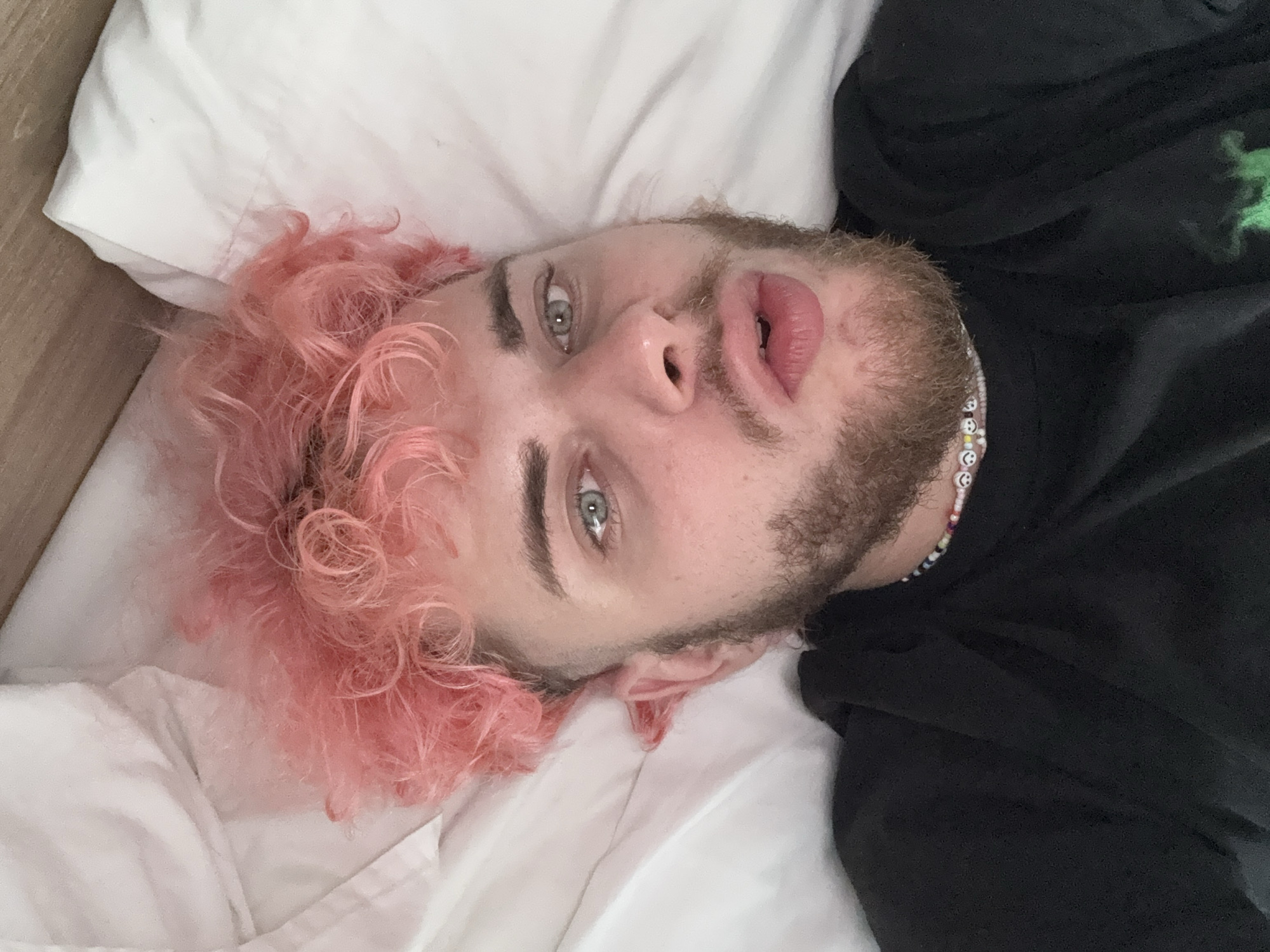 A man with pink, curly hair is lying in a bed with white sheets. He has a beard and fair skin. He is wearing a black shirt and a beaded necklace. His eyes are open, and he is looking at the camera. The image is taken from above and rotated. It is unlikely to be taken in Đà Nẵng, Vietnam.