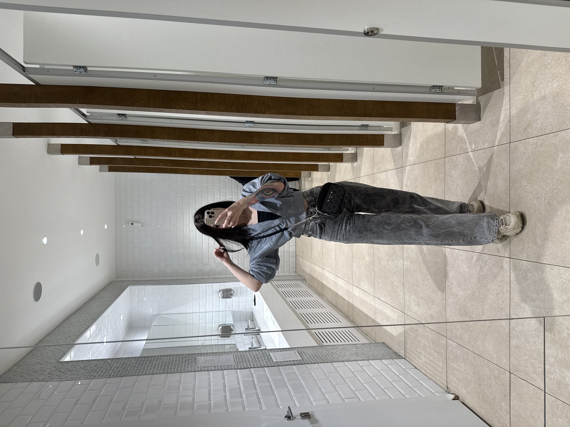 A woman stands in a public restroom, taking a selfie in a mirror. She is wearing a light blue jacket, dark top, gray jeans, and carries a small black bag. The restroom has white subway tiles on the walls and light-colored square floor tiles. A row of stalls with white doors and wooden support beams is visible behind her. The room is well-lit with recessed ceiling lights. There is a mirror spanning the length of the sinks in front of the woman. There is no indication this image was taken in Moscow, Russia.