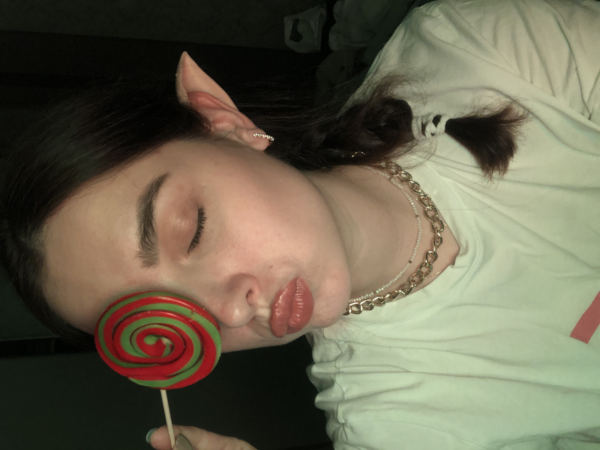 A young Caucasian female with dark hair, styled in a braid, is depicted in a close-up shot, covering her left eye with a red and green swirled lollipop. She has elf-like prosthetic ears, wears a white t-shirt, and is adorned with layered necklaces, one a chunky gold chain and the other a delicate pearl strand. Her lips are pursed, and she has a subtle shimmer on her eyelids. The background is dark and indistinct, suggesting an indoor setting. No specific location in Noviy, Russia, is discernible.