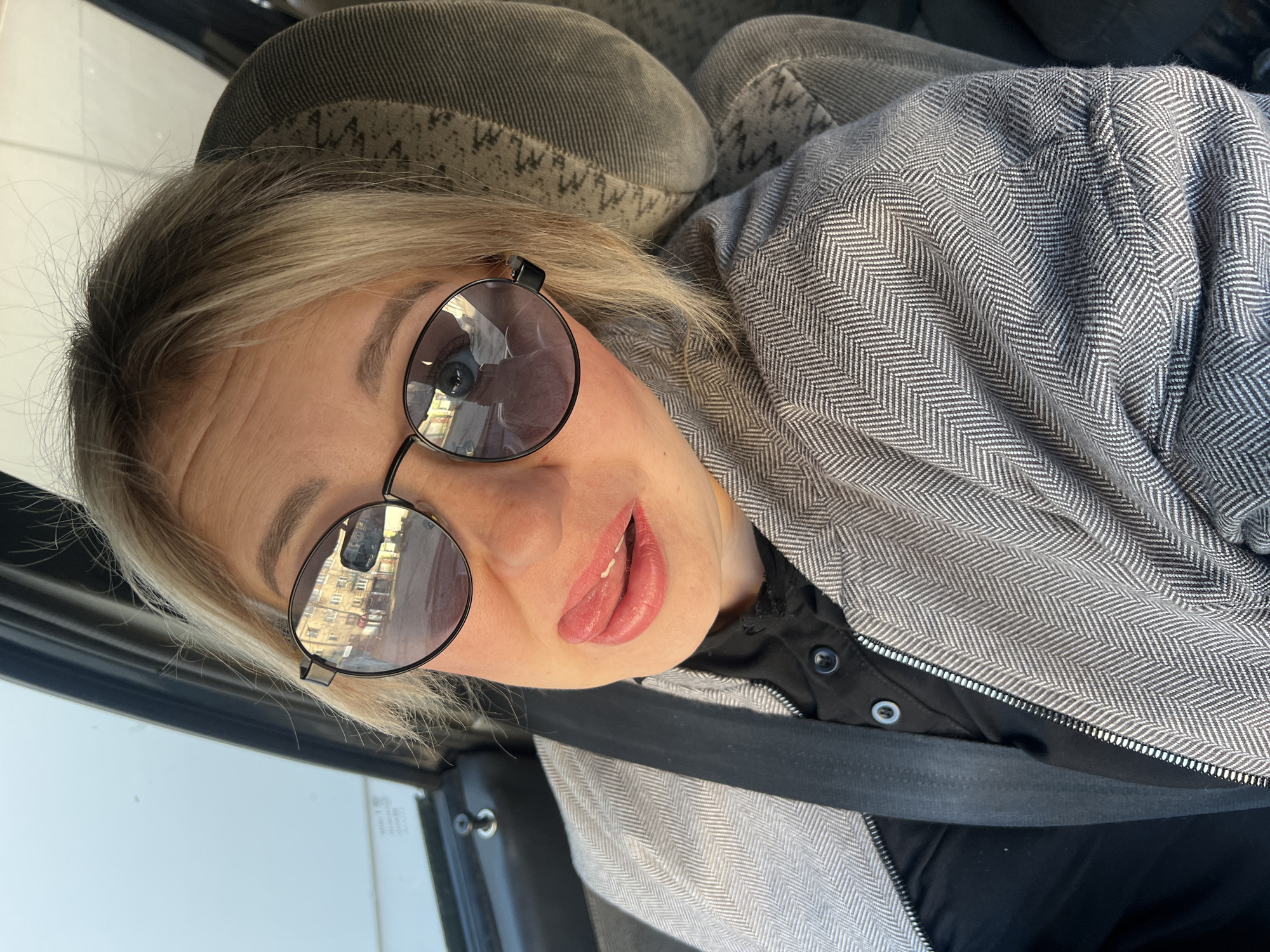 A high-angle selfie captures a fair-skinned woman inside what appears to be a car, tilted to the right. She has light blonde hair, some darker at the roots, and is wearing round, dark-tinted sunglasses with black frames. Her expression is playful, with her mouth slightly open and her tongue peeking out.
She is dressed in a distinctive grey and white herringbone patterned jacket or coat, worn over a black collared shirt visible underneath. A black seatbelt crosses her chest diagonally, indicating she is either a driver or a passenger. The car's upholstered seat, with a textured grey fabric and subtle patterned stitching, is visible behind her head and shoulder.
Reflections in her sunglasses clearly show urban buildings with multiple windows, suggesting the vehicle is moving through or parked in a city environment during the daytime. The bright lighting on the buildings in the reflection confirms it is daytime. The car's window frame is visible on the left side of the image. No visible text can be discerned in the image.