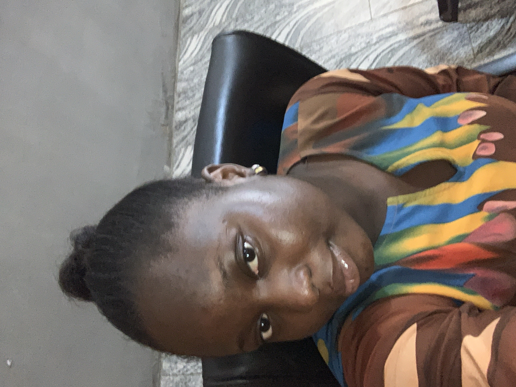 A close-up, medium shot shows a young woman with dark skin, dark eyes, and dark hair pulled back into a bun. She is wearing a multi-colored top with a V-neck. Her left ear, adorned with a small gold stud earring, is visible. The background consists of a dark chair and a wall with a marble pattern. The lighting is warm and suggests an indoor setting, possibly an office or home environment.