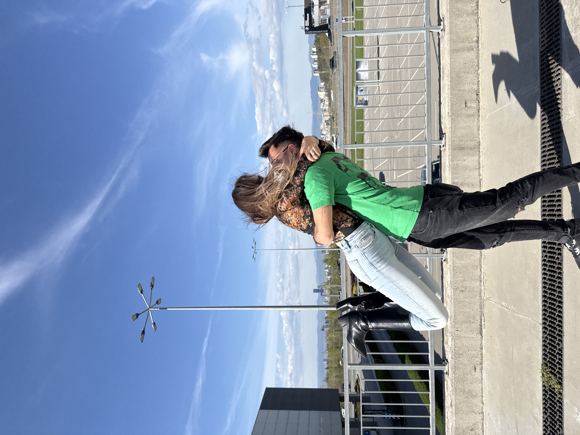 The image captures two individuals, likely a couple, in a close embrace under a clear blue sky in Sofia, Bulgaria. The photo is taken outdoors, seemingly on an elevated urban platform such as a parking garage roof or a similar structure, overlooking a city landscape with distant mountains.
The person on the left, a woman with long, light brown hair, wears a black lace top and light blue jeans. Her arms are wrapped around the other person's neck. The person on the right, a man with dark hair and glasses, is wearing a bright green t-shirt and black jeans, one knee ripped. His arms are around the woman's waist. Their faces are pressed together, suggesting a tender moment or a kiss, with expressions of affection.
The setting is bright, indicating midday or early afternoon, with clear blue skies and only a few wispy clouds. Strong sunlight creates distinct shadows on the concrete ground, including an abstract, jagged shadow resembling a bird or an outstretched hand near a metal drainage grate. A metal railing runs along the edge of the platform. In the background, urban buildings are visible, along with distant mountains under the expansive sky. A tall, multi-lamp street light stands prominently on the left side of the frame. Visible text includes a "BMW" logo on a distant sign and the Cyrillic word "ABTOБУБКА" (Avtobubka or similar) on another sign further back.