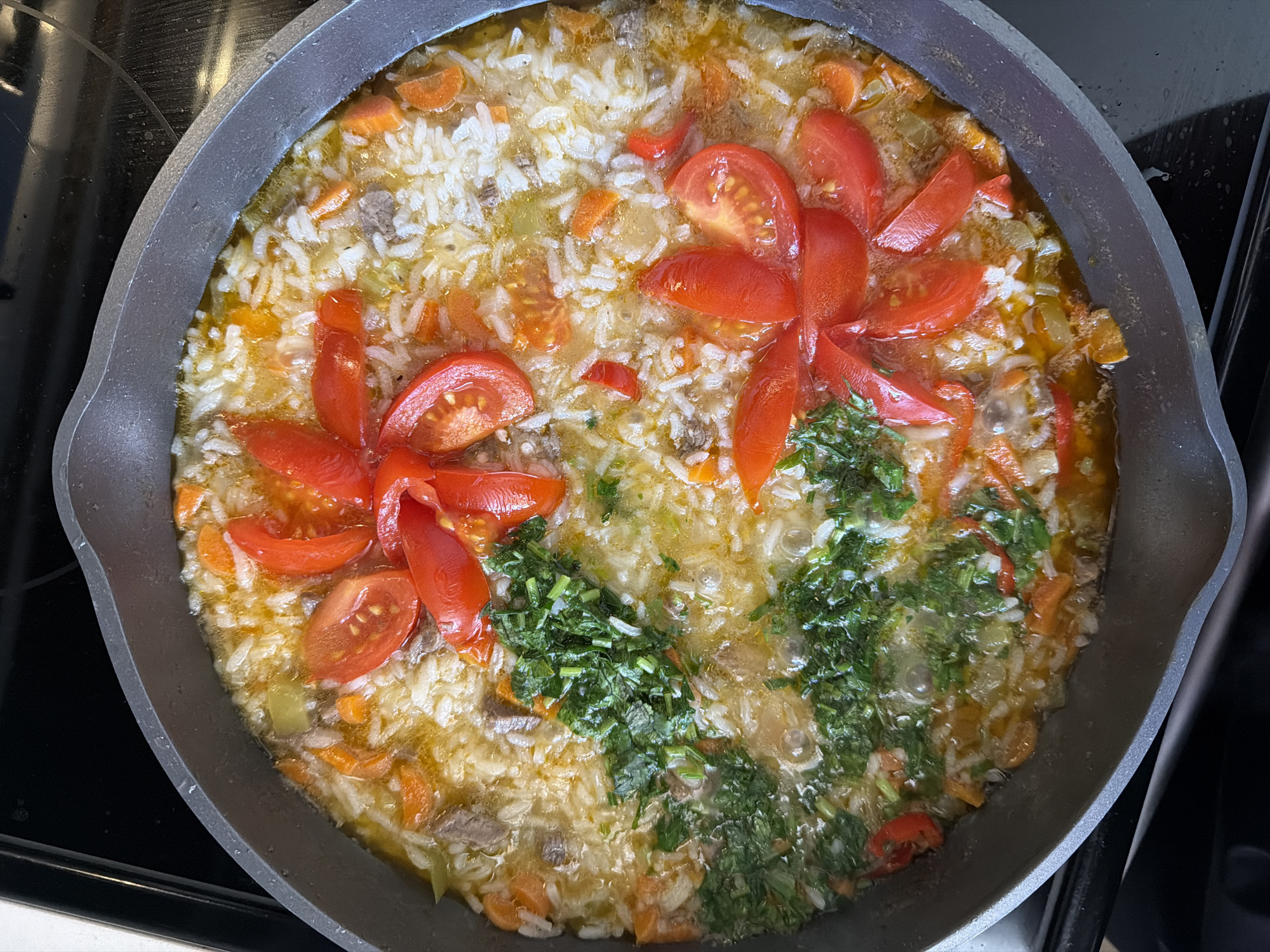 A close-up, overhead shot shows a meal cooking in a dark gray skillet on a black stovetop. The skillet contains rice, diced carrots, small pieces of meat, and what appear to be sliced tomatoes arranged in a flower-like pattern on the surface of the dish. Chopped green herbs are also visible in the center. The food is in a broth that is bubbling, indicating it is actively cooking. The lighting is bright, highlighting the textures and colors of the ingredients. The background is out of focus, but the edge of a white surface is visible to the left, and the black stovetop occupies the rest of the background. There is no indication of a specific location in Miami, United States of America.