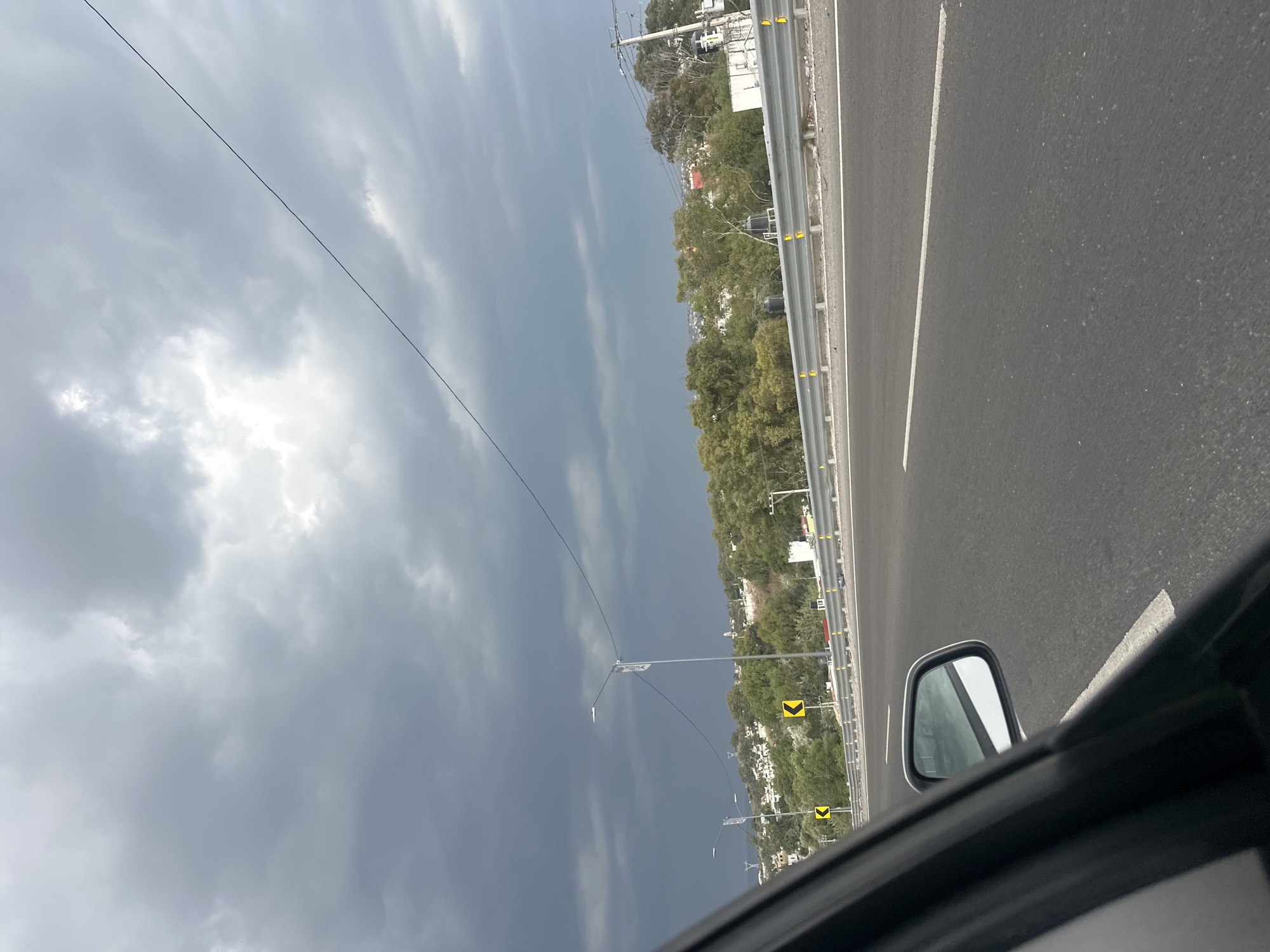 View from inside a moving vehicle on a highway in San Francisco Totimehuacán, Mexico. The road surface is dark gray asphalt with a white lane marker. A metal guardrail runs parallel to the road, interspersed with green trees and buildings. In the distance, utility poles are visible against a cloudy sky. The side mirror of the car is partially visible in the foreground.
