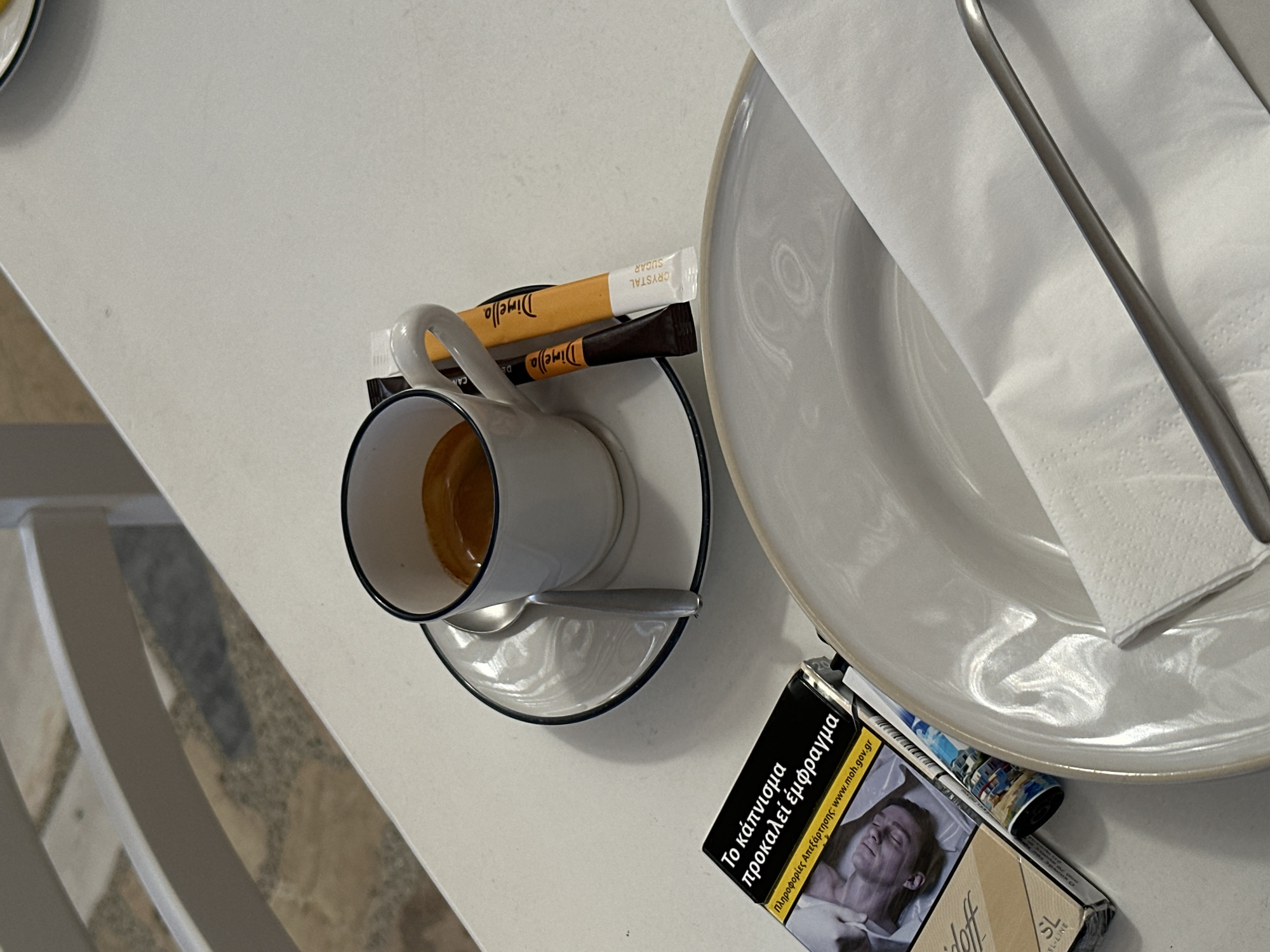 A close-up shot captures a white table with an espresso cup and saucer, two sugar packets, a pack of cigarettes, and a white napkin on a plate. The espresso is dark brown with a thin crema layer. The sugar packets are orange and white, with "Dimello" and "CRYSTAL SUGAR" printed on them. The cigarette pack is black with yellow text and a warning image. A fork rests on the napkin. The background shows white chairs and a tiled floor, suggestive of a cafe or restaurant setting in Ano Mera, Greece. The lighting is bright, casting soft shadows on the table.