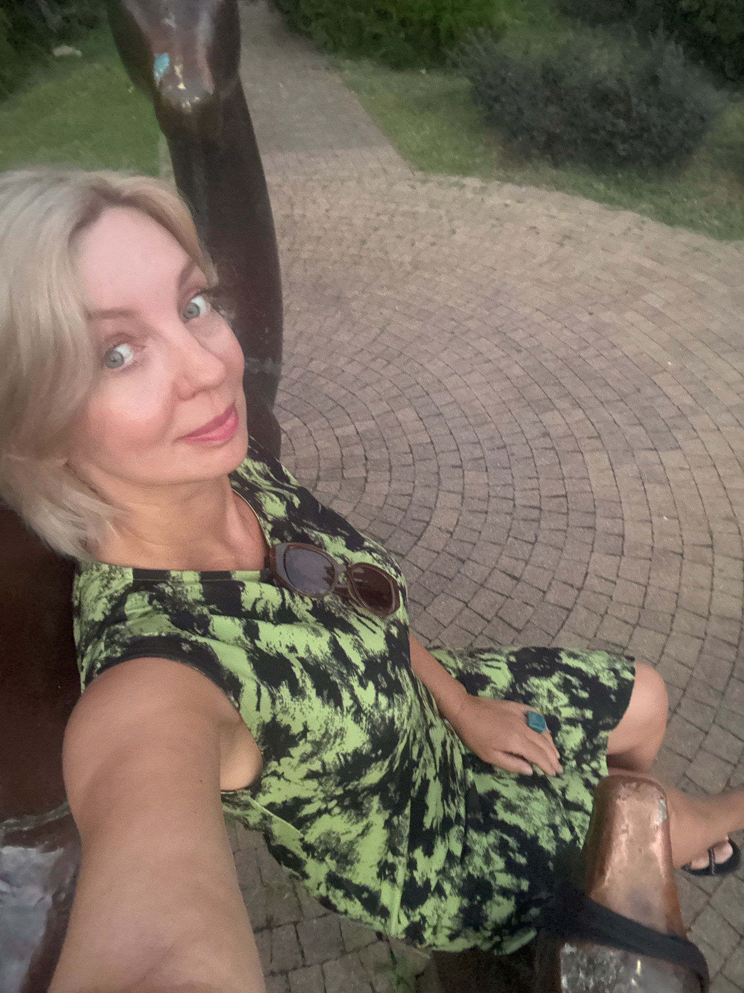 A woman with blonde hair and blue eyes, wearing a green and black patterned dress, sits outdoors on a paved path. She is looking at the camera and smiling slightly. Sunglasses are resting on her chest. She is wearing a large turquoise ring on her right hand. A dark bronze sculpture is partially visible to her left. The background consists of a paved walkway with a curved pattern, grass, and bushes. This scene is located in the city of Sochi, Russia.