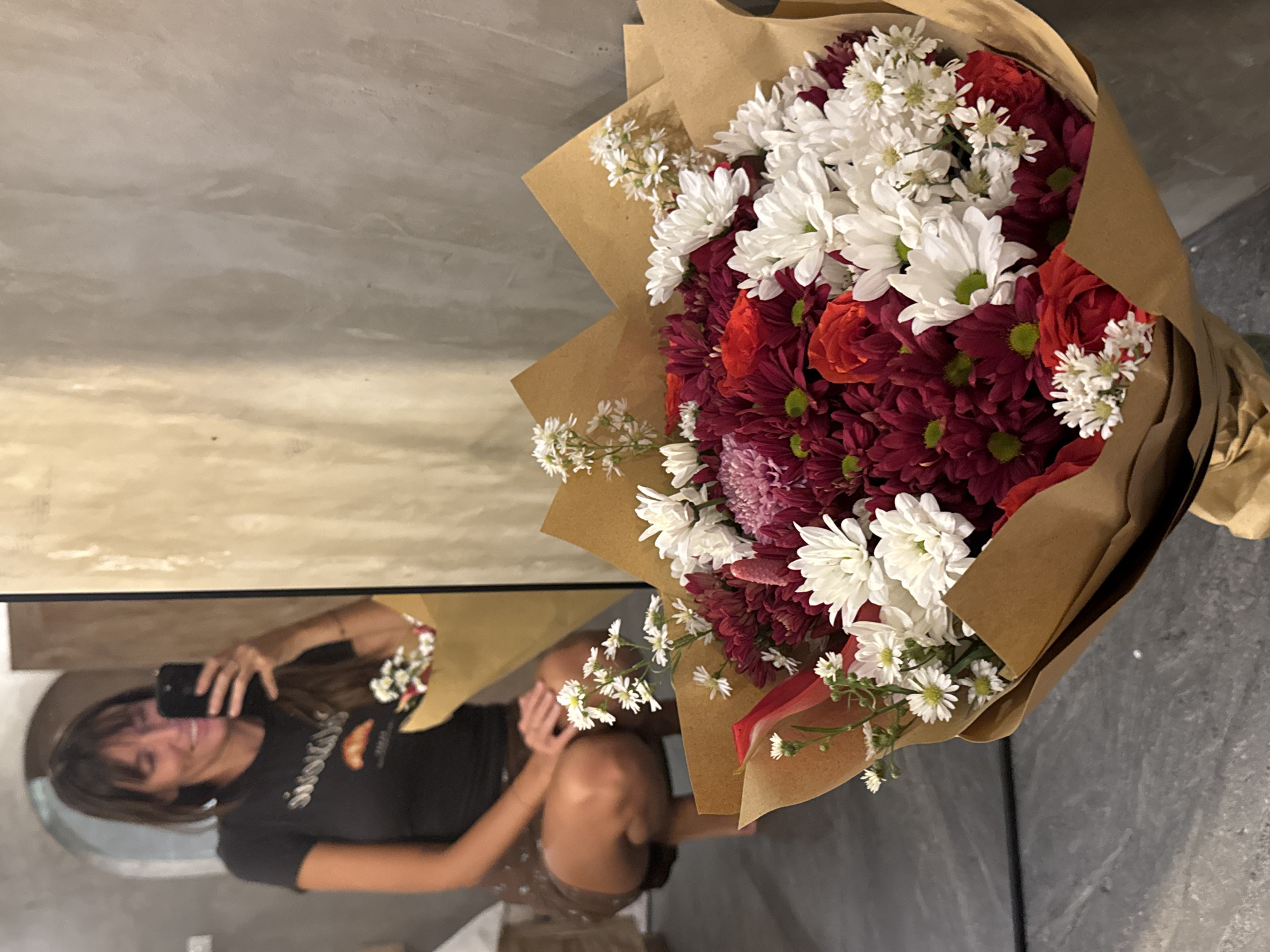 A woman is taking a mirror selfie with a bouquet of flowers. The bouquet is large, wrapped in brown paper, and features red, burgundy, and white flowers. The woman is reflected in the mirror, holding a cell phone to take the picture. She is wearing a dark-colored t-shirt and shorts. The mirror reflects a room with light-colored walls. The location is North Kuta, Indonesia.
