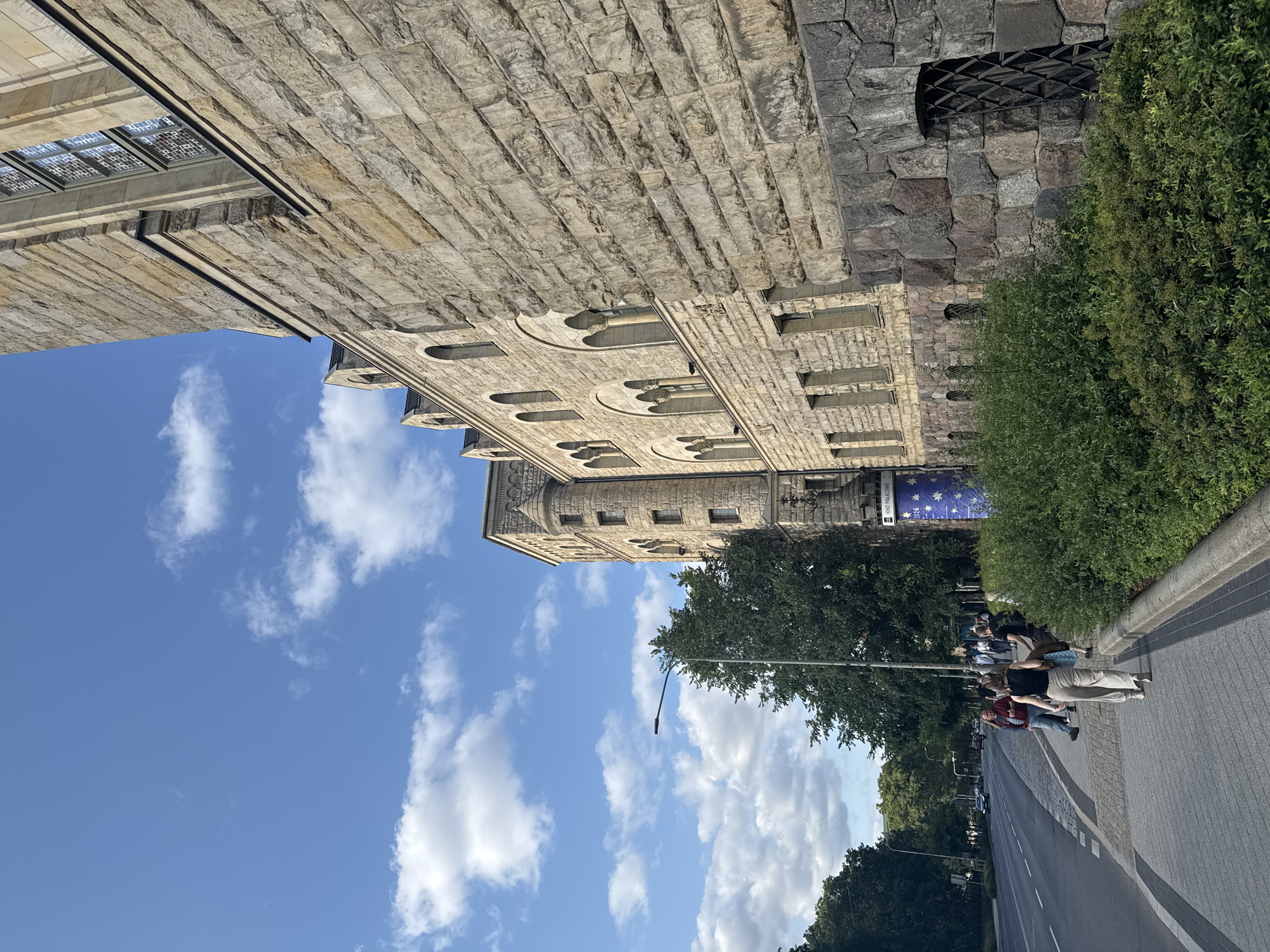 The image depicts a street scene in Poznan, Poland, with a prominent stone building on the right side. The building features a weathered stone facade, arched windows, and a tower-like structure. A sidewalk runs along the building, bordered by a street on the left. Several pedestrians are walking on the sidewalk, some carrying bags. Lush green trees and bushes are visible along the sidewalk and in the background. The sky is blue with scattered white clouds. A street lamp is positioned on the sidewalk. A sign with the text "KINO PAŁACOWE" and stars is attached to the building. The overall scene appears to be a daytime view of an urban environment.