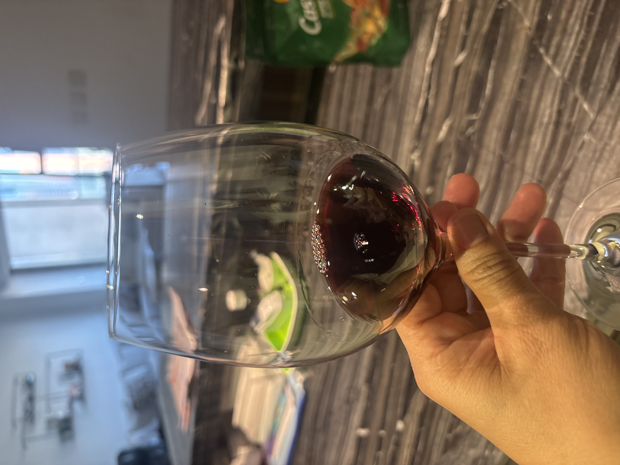 A close-up, first-person perspective shot shows a hand holding a stemmed wine glass filled with red wine. The glass has small bubbles on the surface of the wine. The background is blurred and appears to be an indoor setting, possibly a kitchen or living area, with a marble countertop. On the countertop, a green bag with the word "Castello" is visible. The overall scene suggests a relaxed, indoor environment. The image does not provide any information about a specific location in Dubai, United Arab Emirates.