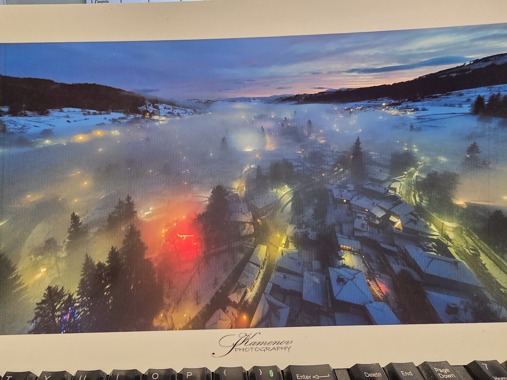 This media file displays a panoramic photograph of a snow-covered settlement nestled in a valley, likely at dusk or dawn. The scene is enveloped in a soft, ethereal mist or fog, which diffuses the numerous artificial lights scattered throughout the town.
**Subjects:** The primary subjects are the snow-covered buildings, houses, and trees, forming a quaint village or small town. Mountains or hills flank both sides of the valley, also blanketed in snow. The sky above transitions from deep blue to soft pink and orange hues, indicative of twilight.
**Activity or event taking place:** The photograph captures a serene winter evening or early morning scene, with the town's lights illuminating the mist-laden landscape. The presence of colorful lights, particularly a prominent red glow on the left and some blue and red lights on trees, suggests a festive or holiday atmosphere.
**Notable details:**
- **Time of day:** The sky's colors and the illuminated artificial lights strongly suggest either twilight (dusk or dawn).
- **Weather:** Heavy snowfall is evident, covering roofs and ground, and a significant amount of mist or fog hangs low, creating a dreamy effect.
- **Location cues:** The mountainous terrain and dense clusters of pine-like trees point to a location in a mountain region, possibly a ski resort or an alpine village.
- **Emotions:** The image evokes feelings of peace, tranquility, magic, and coziness, typical of a winter wonderland scene.
- **Lighting:** Streetlights, house lights, and decorative lights cast warm yellow, orange, and red glows, creating a striking contrast against the cool blue and white tones of the snow and sky. The mist amplifies the glow of these lights.
**Visible text:** At the bottom center of the image, the text "Kamenov PHOTOGRAPHY" is clearly visible, serving as a signature or watermark. Additionally, partially visible text at the very top of the original image reads "Георги" (Georgi in Cyrillic).