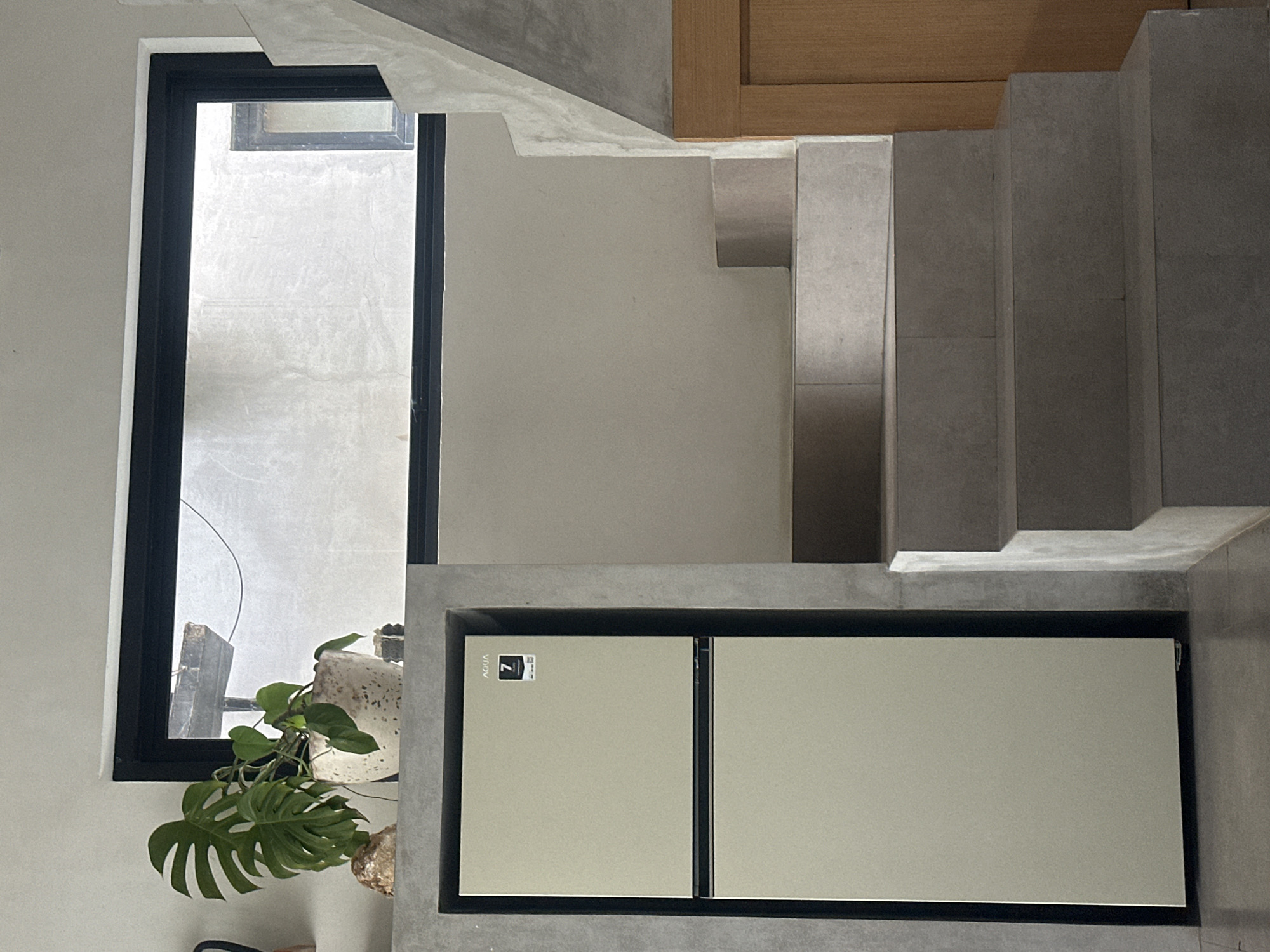 The image presents an interior space, possibly a residential dwelling, located in Mengwi, Indonesia. A dark-framed window on the left provides a view of an outdoor area, revealing a textured, light-colored wall and some greenery. A plant with large, split leaves is situated near the window, inside the building. Adjacent to the window, the wall is painted in a neutral tone. To the right of the wall, a flight of gray concrete stairs ascends upwards. Under the stairs, there is a built-in cabinet or appliance, possibly a refrigerator, which is rectangular with two doors and a dark frame. The surrounding structure and walls exhibit a concrete-like texture.