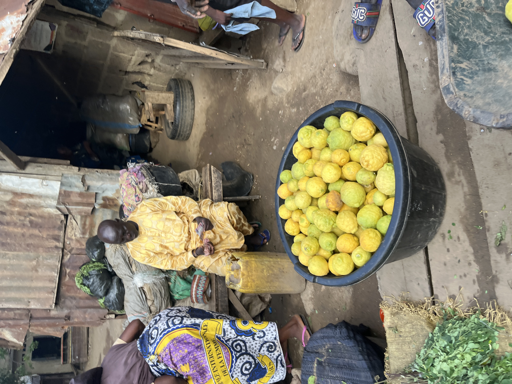 A man wearing a yellow traditional outfit sits on a wooden bench in what appears to be an outdoor market stall in Rafin Par, Nigeria. He holds a small bundle of items in his hands. To his left, a woman in a colorful patterned skirt stands, her back to the camera. In front of them, a large black tub is filled with numerous yellow-green citrus fruits, likely lemons or limes. A tall, yellow plastic container with some liquid is positioned to the right of the fruit tub. The background consists of a rustic structure made of corrugated metal and mud brick, with various sacks and bundles of goods visible. The ground is dirt and appears damp. To the right of the image, another person's legs and feet wearing sandals are visible, along with a blue towel. A pile of green leafy vegetables is in the lower left corner, next to a woven mat. The scene depicts a typical market setting with a focus on produce sales.