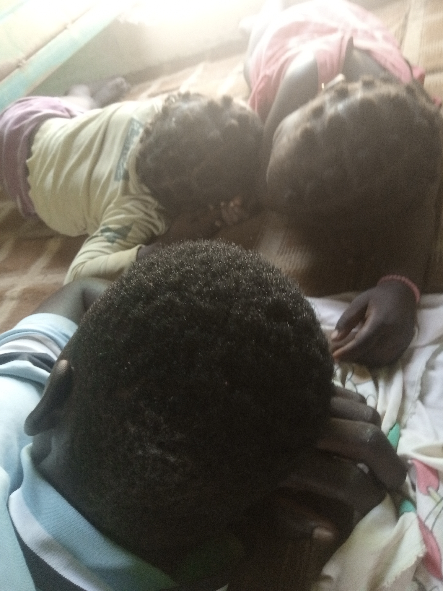 In a dimly lit room in Langtang, Nigeria, three individuals appear to be sleeping or resting on a surface covered with a patterned cloth. The person in the foreground, seen from the back of the head, is wearing a light blue and white striped collar shirt. Their hand is raised and placed over their face. To their left and slightly further back, another person is lying on their side, facing away from the camera. They are wearing a light yellow long-sleeved shirt and what appears to be purple shorts or a skirt. Further behind them, the back of a third person's head is visible, wearing a pink garment. The overall impression is one of a domestic or communal resting space, with soft lighting and a casual arrangement of individuals.