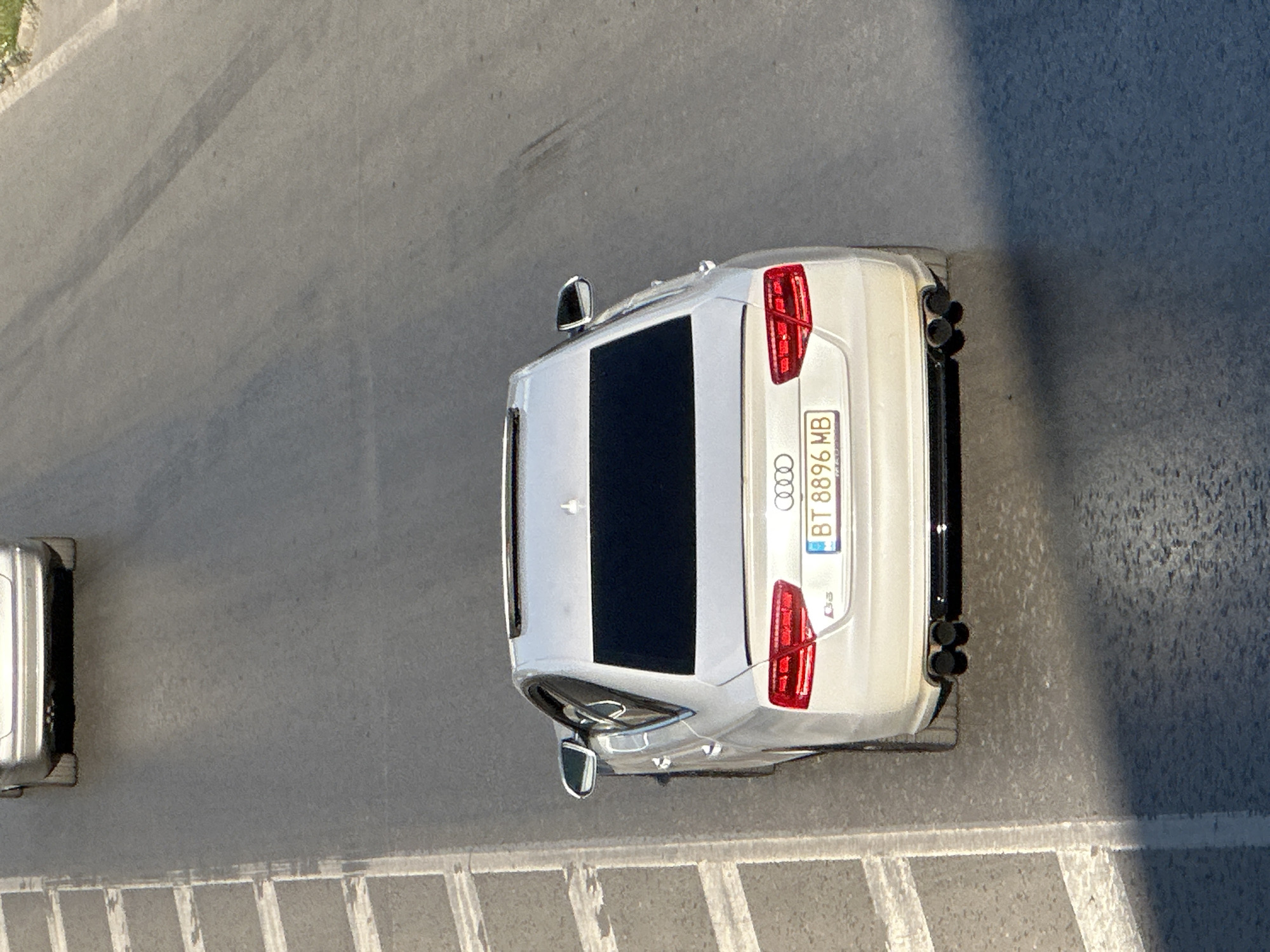 This media file contains an overhead shot of a white Audi parked on a grey asphalt road, likely in Veliko Tarnovo, Bulgaria, given the license plate's format (BT 8896 MB). The car is the primary subject, seen from directly above. It's a modern sedan, with its rear facing the viewer. Notable details include its quadruple exhaust pipes, tinted windows, and sleek design. The license plate is clearly visible, displaying "BT 8896 MB" in white lettering on a blue and yellow background, which is consistent with Bulgarian license plates.
The scene appears to be an outdoor setting, possibly a street or a parking area. The road surface shows some texture and there are faint tire marks, suggesting recent activity. To the left of the Audi, another vehicle, a silver van or small truck, is partially visible, parked parallel to the road. A thin white line, possibly a road marking or curb, is visible on the far left edge of the image. A long, dark shadow stretches across the right side of the asphalt, indicating it's likely daytime, with the sun casting a distinct shadow from an unseen object. The overall lighting suggests a bright day.
There are no people visible in the frame. The activity depicted is simply a parked car on a road. The setting appears to be urban or suburban, with the texture of the asphalt and the presence of multiple vehicles suggesting a developed area. The image is captured with a high angle, providing a clear, unobstructed view of the car and its surroundings.