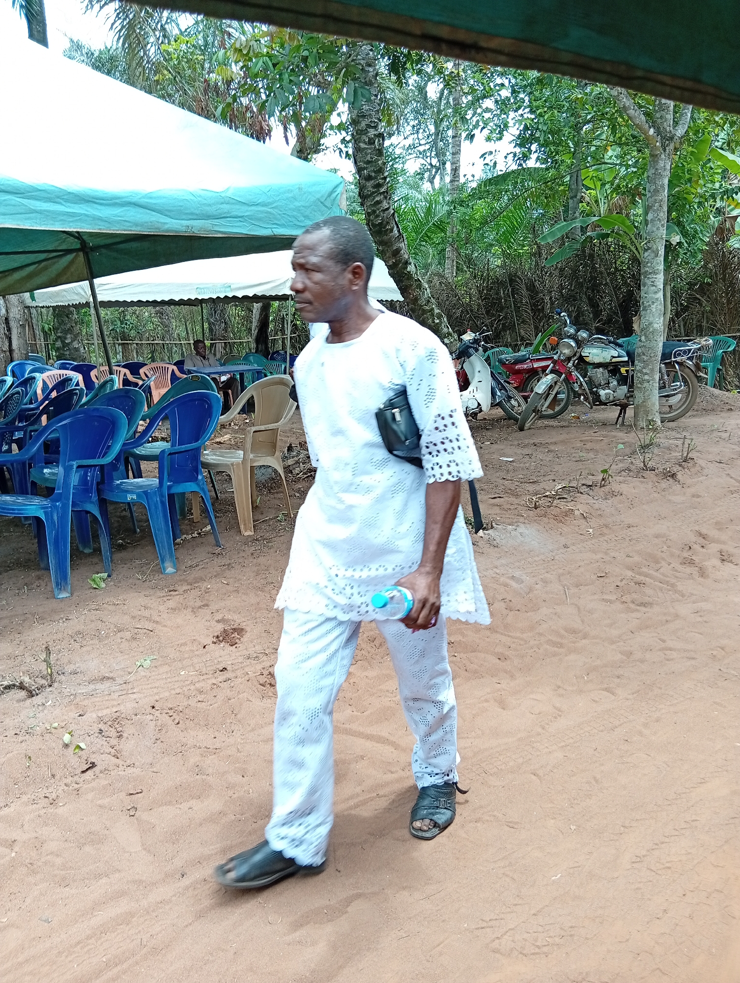 The image captures an outdoor scene, likely a temporary gathering or event in Umundugba, Nigeria. The ground is a light brown dirt or sand, showing some footprints and tire tracks. In the foreground, a man, appearing to be of African descent, walks from left to right. He is dressed in a clean, white, two-piece traditional or semi-traditional outfit, featuring intricate lace-like cutouts on the sleeves and the bottom edges of both his long top and trousers. He wears dark open-toed sandals and carries a small black crossbody bag over his left shoulder. In his right hand, he holds a clear plastic water bottle with a light blue cap. His gaze is directed slightly ahead and to his right, with a neutral expression.
The background features several elements indicating a communal setup. To the left, under a large canopy tent with a white top and light blue underskirt, numerous plastic chairs are arranged. The chairs are mostly blue, with some beige and orange ones, suggesting seating for attendees. Further back, beneath another tent structure, a person can be vaguely seen seated at a table. On the right side of the frame, behind the walking man, two motorcycles are parked on the dirt, one red and one with white and dark colors. Lush green foliage and various trees, including palm trees, form the backdrop, indicating a natural, possibly rural or semi-urban environment. The lighting suggests it is daytime. No visible text or prominent branding is discernible in the image.