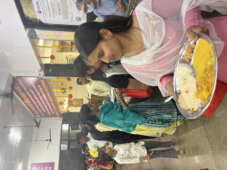 The image shows a group of people in a dining hall in Panchkula, India, located at 30.723 latitude and 76.862 longitude. The people are standing in a line, waiting to receive their food. A young woman in a pink and white salwar kameez is holding a tray of food, which includes a roti, rice, and a yellow curry. The woman in front of her is wearing a blue and white salwar kameez. There are several other people in the line, including a young girl in a white shirt and jeans, a man in a black shirt, and several other women. The dining hall is decorated with garlands of marigold flowers. There is a sign on the wall that reads "Shradhanjali Bhawan Veg.  Kitchen (Meal Kary).". There is also a certificate from the "TCCPCL" organization, which certifies the hall as being "ISO 9001:2015" compliant.  The room is well-lit and there is an overhead fan in the ceiling. The floor is tiled.  The woman in the pink salwar kameez is looking towards the camera.  The image captures a scene of people waiting for their food in a bustling dining hall.