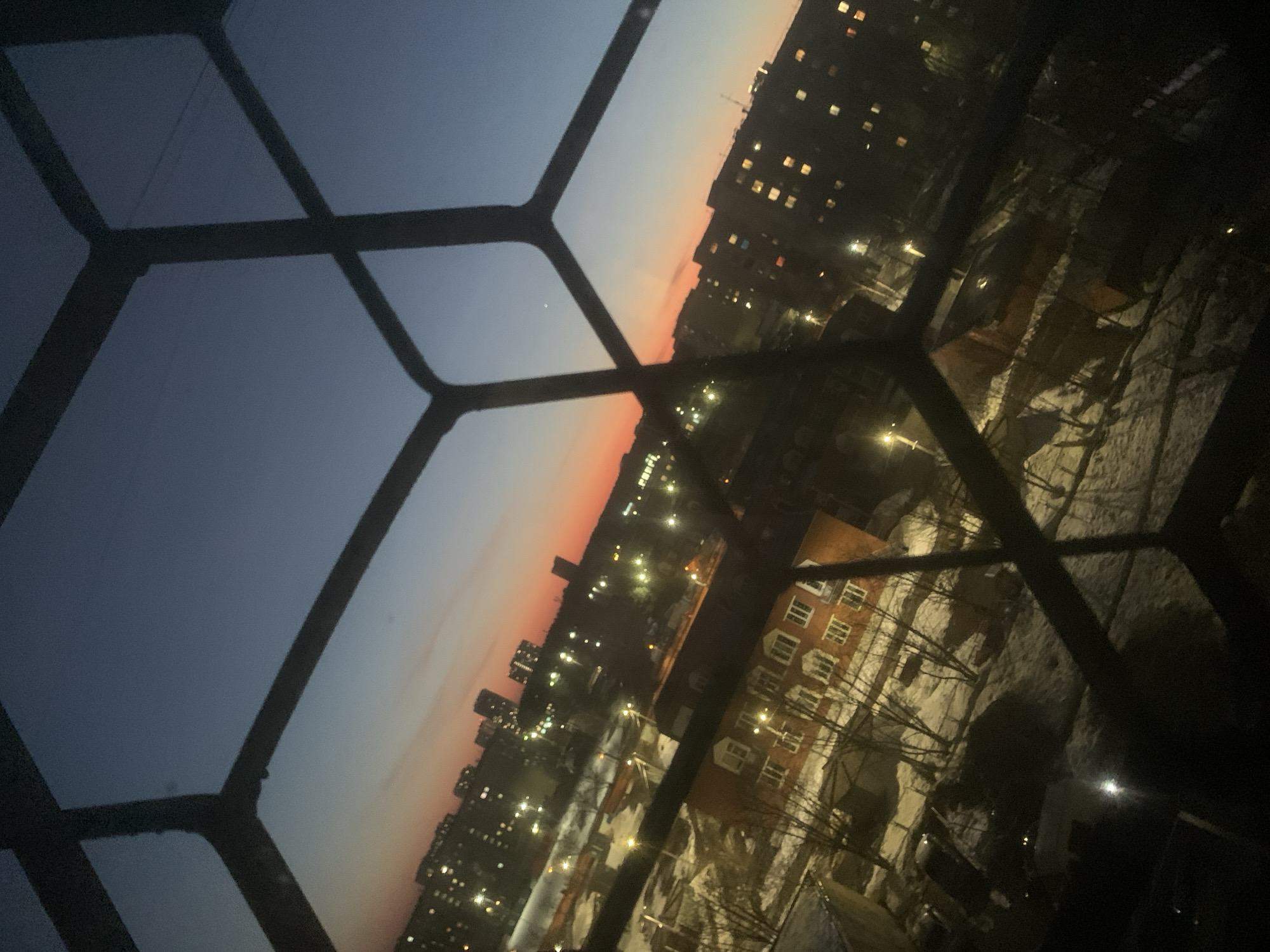 The image shows a nighttime view from a window with a decorative frame. Outside, a cityscape is visible under a sky transitioning from dark blue to a faint orange glow on the horizon, indicating either sunset or sunrise. Buildings of varying heights are illuminated with numerous lights. In the foreground, a residential area is covered in snow with visible streetlights. The architectural style of the buildings suggests a location in Izhevsk, Russia.