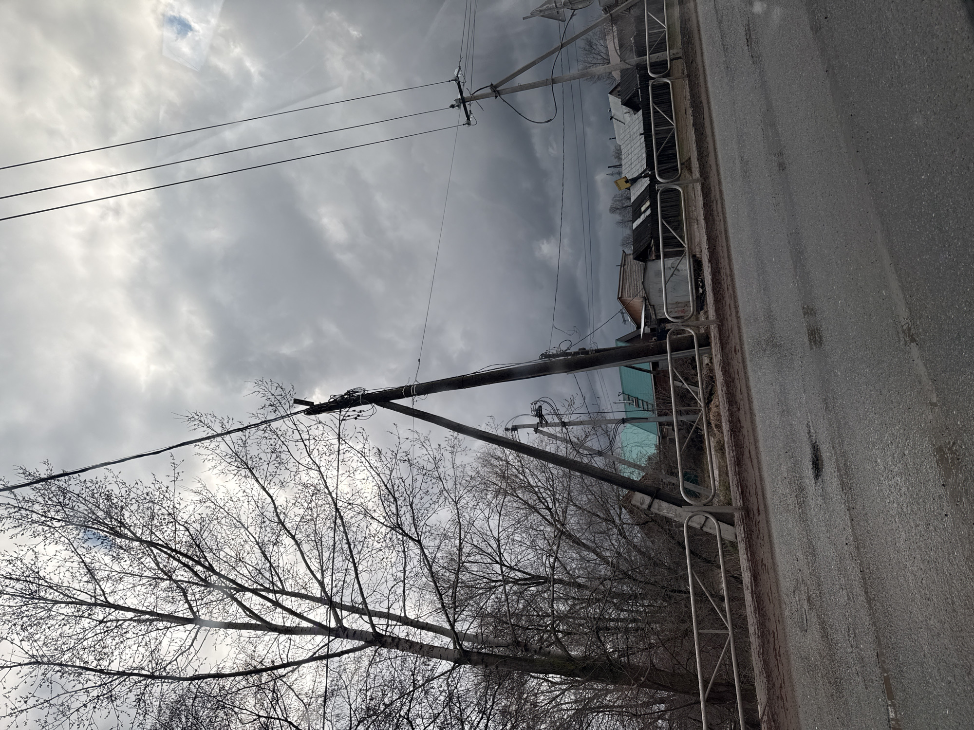 This daytime photograph captures a street scene in Chaikovsky, Russia, on an overcast day. The image is taken from a slightly low perspective, looking down a street and towards the horizon.
In the foreground, a paved asphalt road occupies the bottom and right side of the frame. A simple metal guardrail or fence, composed of vertical posts and rounded top sections, runs along the edge of the road.
The midground is dominated by several utility poles, appearing to be made of wood, which stand tall with numerous crisscrossing power and communication lines. These lines extend across the sky, connecting the poles. To the left, a large deciduous tree, completely bare of leaves, fills a significant portion of the scene, its dark branches silhouetted against the bright, cloudy sky. More bare trees are visible further back.
In the background, beyond the poles and trees, various modest buildings, possibly houses or sheds, can be seen with their roofs and upper structures. Some exhibit light-colored siding or painted surfaces.
The sky is heavily overcast with dense, gray clouds, though some lighter areas suggest varying cloud thickness. The overall lighting is diffuse, creating a muted and somewhat somber atmosphere typical of a cloudy winter or early spring day. There are no people visible, and no discernible text appears in the image.