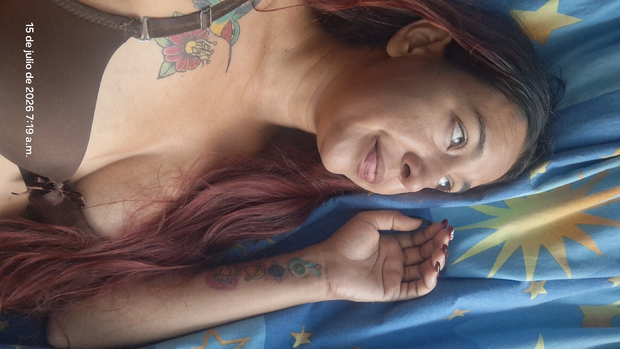 A woman with long, reddish-brown hair is lying on her back, looking up and to her left. She is wearing a brown bra and has a tattoo of a hummingbird and flowers on her left shoulder and chest. A tattoo of celestial bodies is visible on her right wrist. She is lying on a blue fabric with yellow star patterns. The timestamp "15 de julio de 2026 7:19 a.m." is superimposed on the bottom left corner of the image. The scene appears to be indoors, likely in a bedroom. The location is identified as Vallecito, Venezuela.