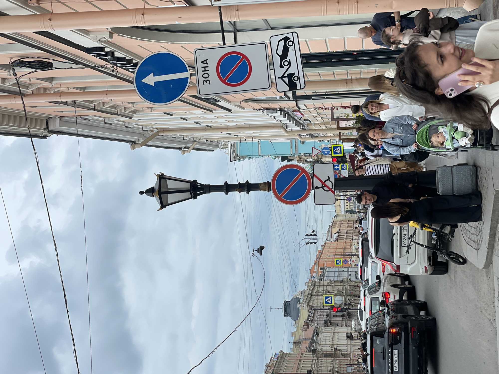 A street scene in Saint Petersburg, Russia, on a cloudy day. Several cars are stopped in traffic, including a black Haval and a white Volvo. Pedestrians walk along the sidewalk, some pushing strollers or carrying bags. Several traffic signs are visible, including a "No Stopping" sign and a sign indicating a towing zone. The buildings lining the street are tall and narrow, with ornate facades. The sky is overcast with gray clouds.