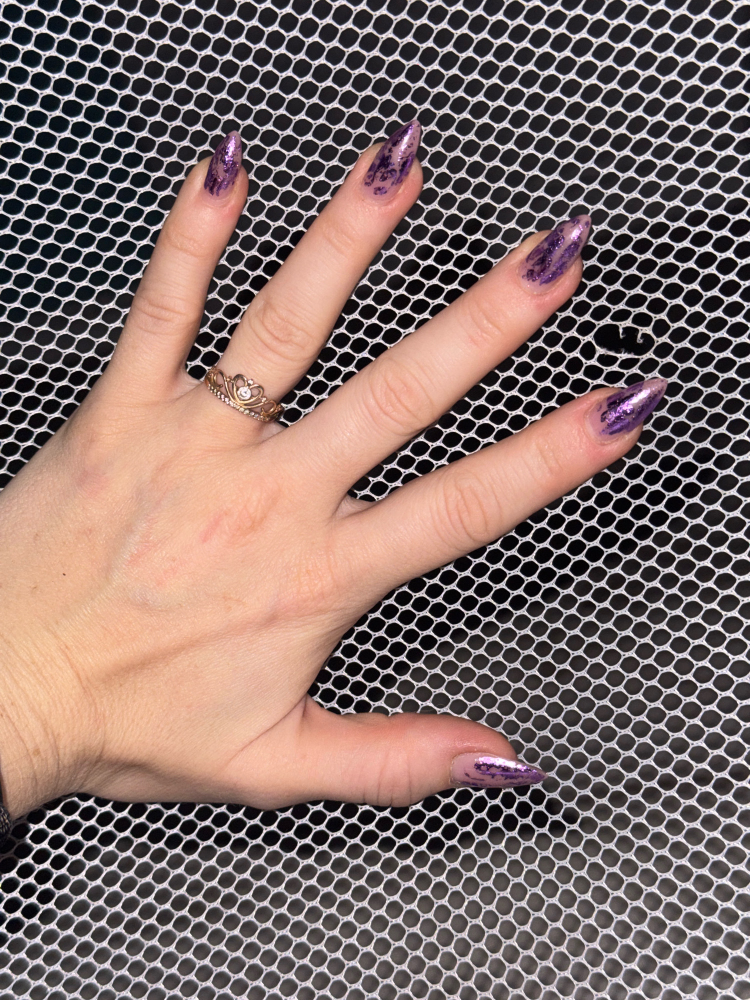 In the city of Rusalevka, Russia, a person's left hand is prominently displayed against a black background with a white mesh overlay. The hand, with its fingers extended and slightly splayed, showcases a set of manicured nails. The nails are long and almond-shaped, painted with a sparkling purple polish that has a textured, holographic effect. The pinky finger nail appears to have a clear base coat with flecks of purple glitter. A delicate gold ring, designed to resemble a crown with small, clear stones embedded within it, adorns the ring finger. The skin tone of the hand is fair, and there are a few faint red marks visible on the back of the hand, possibly from minor abrasions. The overall lighting is bright, casting subtle shadows and highlighting the texture of the mesh and the shimmer of the nail polish.