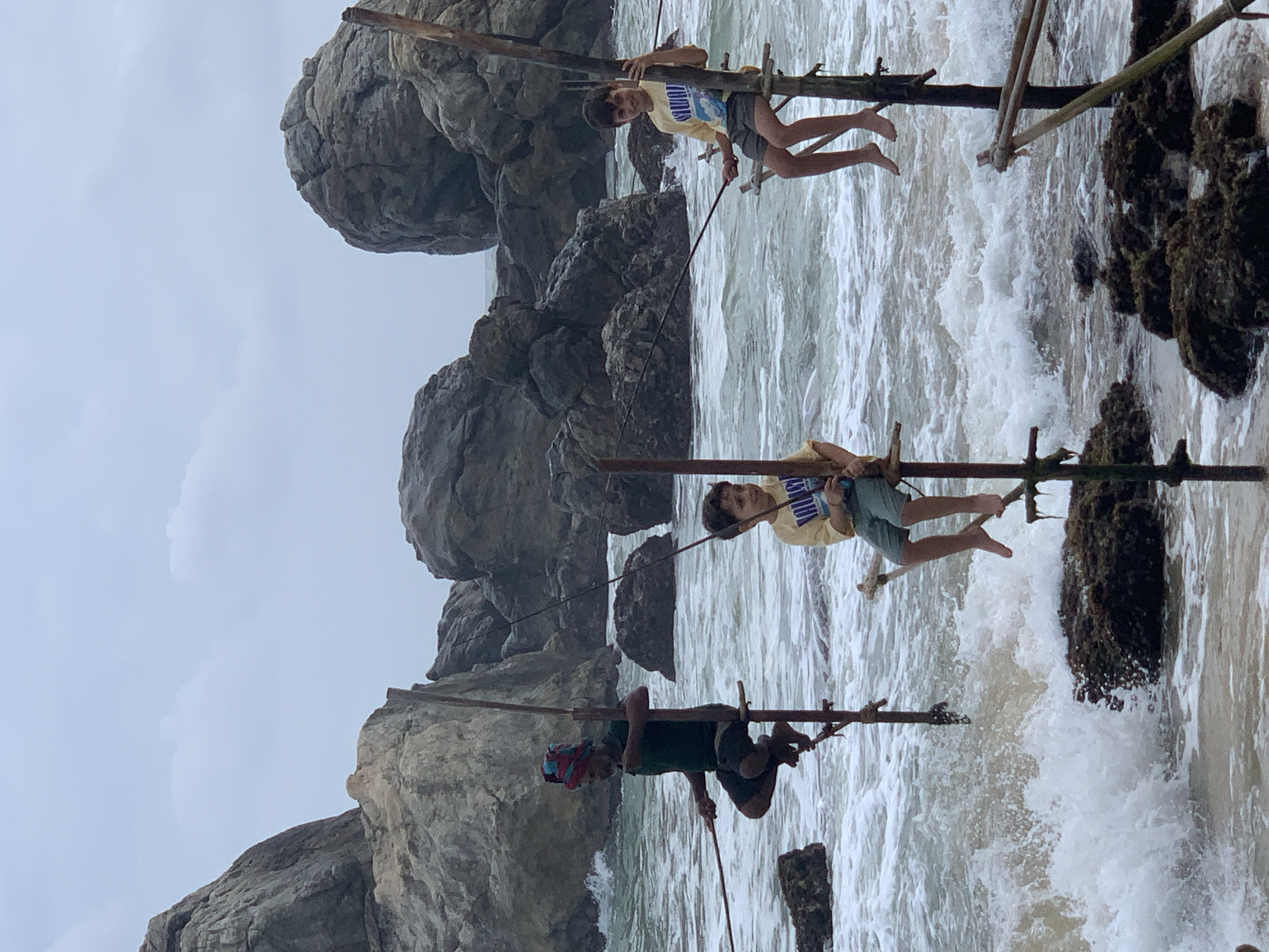 The image shows three people engaged in stilt fishing on a rocky coastline in Kathaluwa, Sri Lanka. In the foreground, a man in a green shirt and a colorful turban is perched on a wooden stilt, holding a fishing rod. To his right, two young boys are also on stilts, one in a yellow shirt with blue graphics and the other in a similar yellow shirt. The boys appear to be observing or participating in the fishing activity. The background features large, weathered rocks and the ocean with white-capped waves crashing against the shore. The sky is overcast, suggesting a cloudy day. The stilt fishing technique, a traditional method in Sri Lanka, involves fishermen balancing on a narrow perch tied to a vertical pole, with their fishing rods extended into the water.