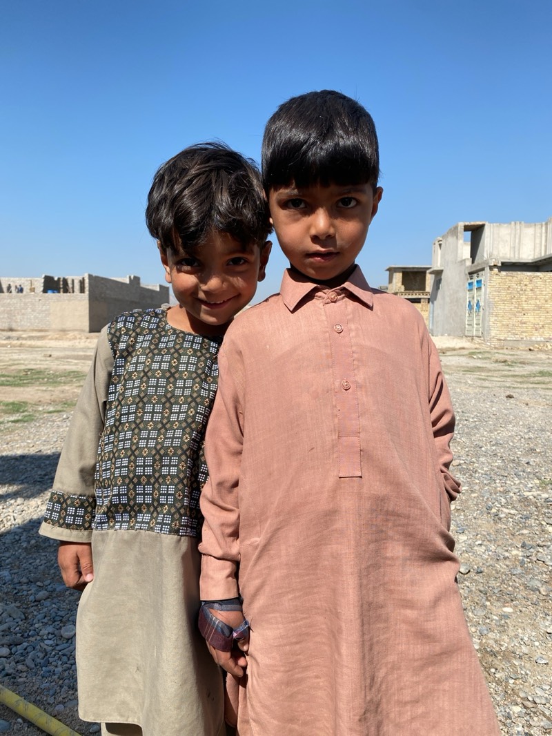 Two young boys stand close together in what appears to be Kandahar, Afghanistan, based on the traditional clothing and the architectural style of the buildings in the background. The boy on the left has dark, wavy hair and wears a tan and patterned shirt. He is smiling slightly. The boy on the right has straight, dark hair and wears a salmon-colored tunic. Both appear to be wearing traditional Afghan clothing. The ground is rocky and dusty, with unfinished or partially constructed buildings visible in the distance under a clear, blue sky. The scene suggests a rural or developing area within Kandahar.