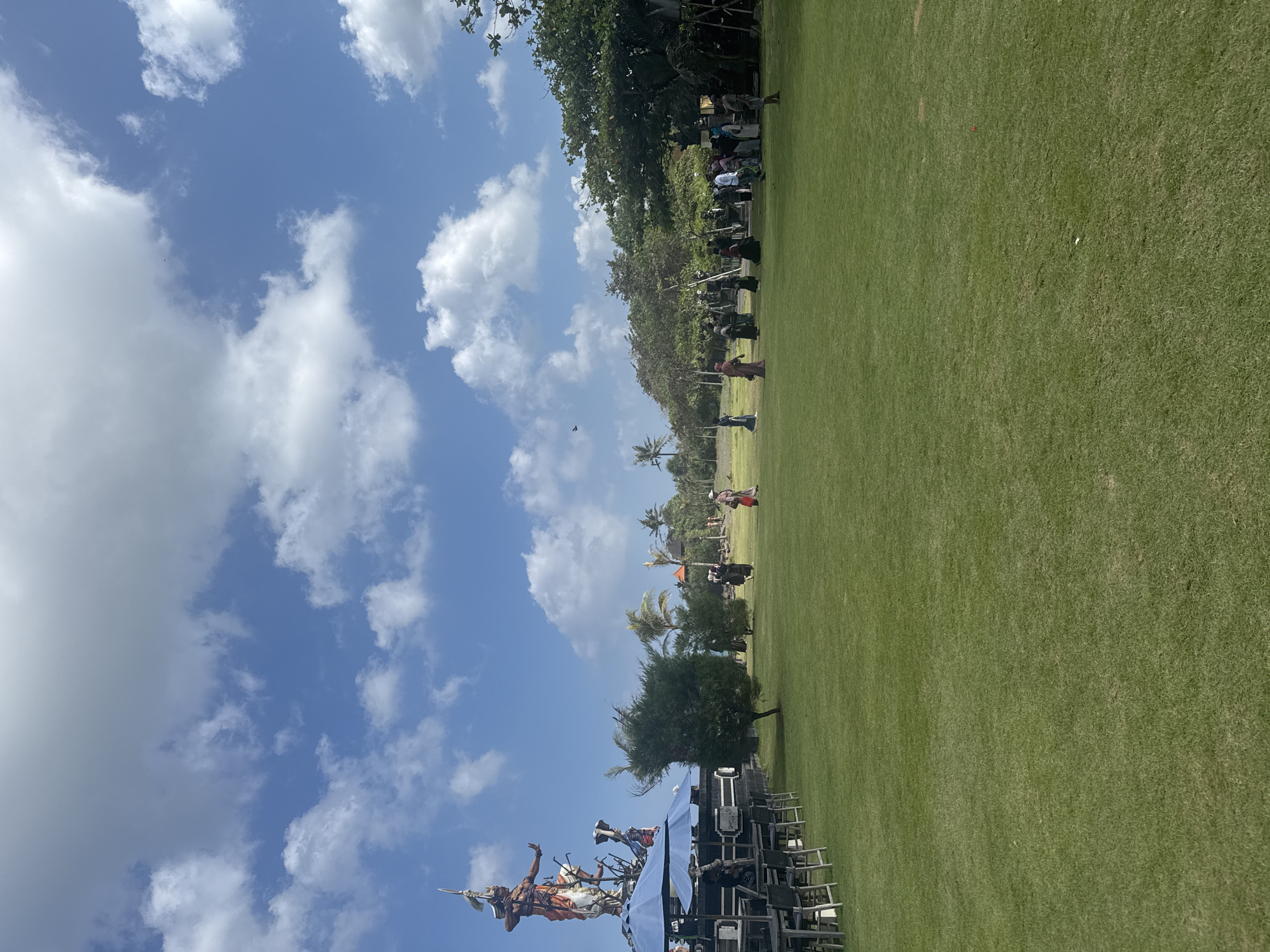 An outdoor scene displays a large grassy field under a partly cloudy blue sky. To the left, a tall, ornate statue of a figure with outstretched arms stands against the sky, accompanied by smaller figures and ornate structures. In front of the statue, several tables and chairs are set up under blue umbrellas, suggesting a seating or dining area. A group of people, some in traditional attire, are scattered across the field, appearing to be observing or participating in an event. Trees and foliage line the background, with a few buildings visible amidst the greenery. The overall impression is of a cultural or recreational gathering in a tropical or subtropical setting.