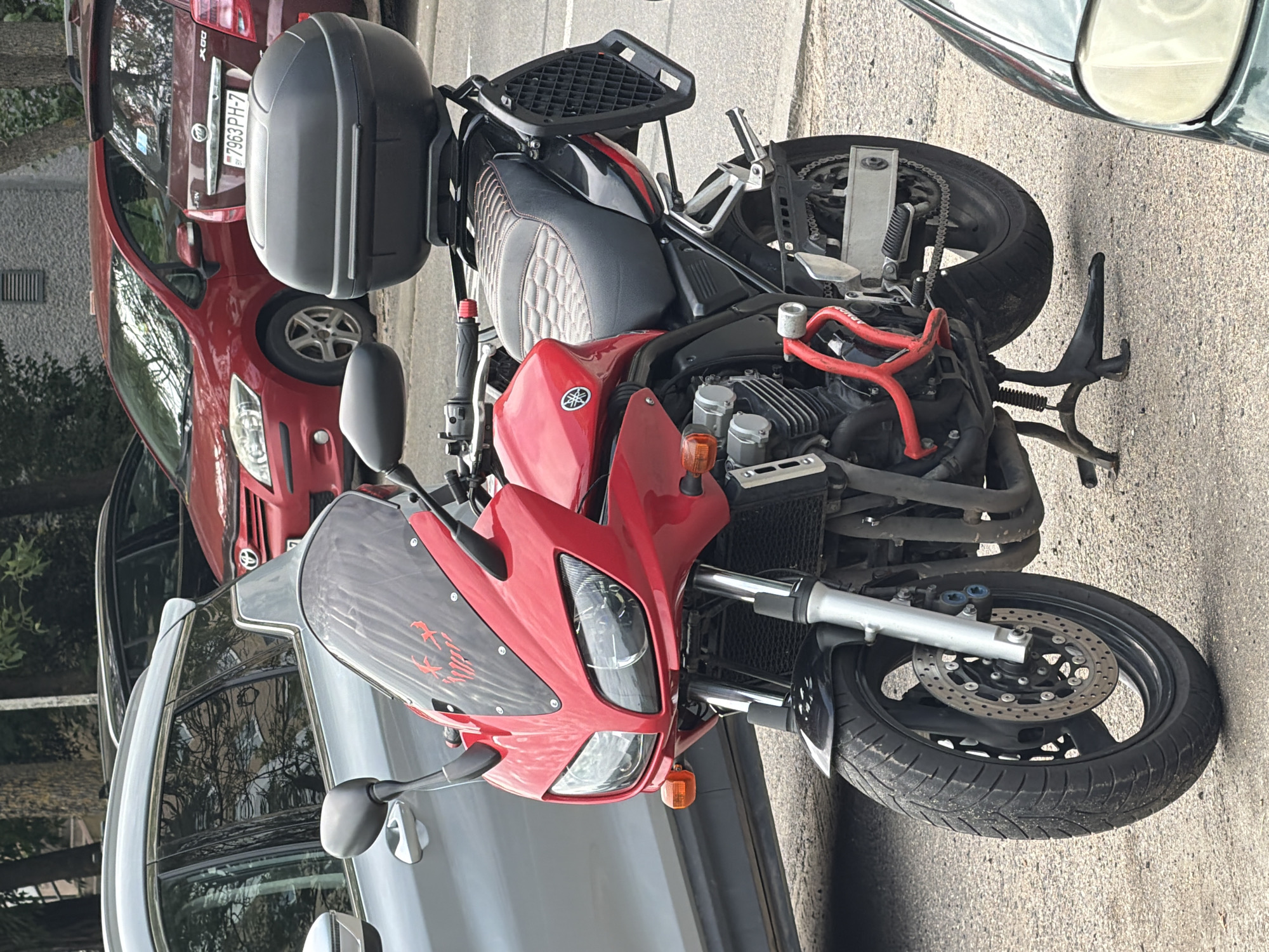 A red Yamaha motorcycle is parked on a paved surface in front of several cars, including a silver SUV and a red sedan. The motorcycle has a black top box and a black padded seat. The red sedan has a Belarusian license plate that reads "7963 PH-7". The scene appears to be a street or parking area in Minsk, Belarus.