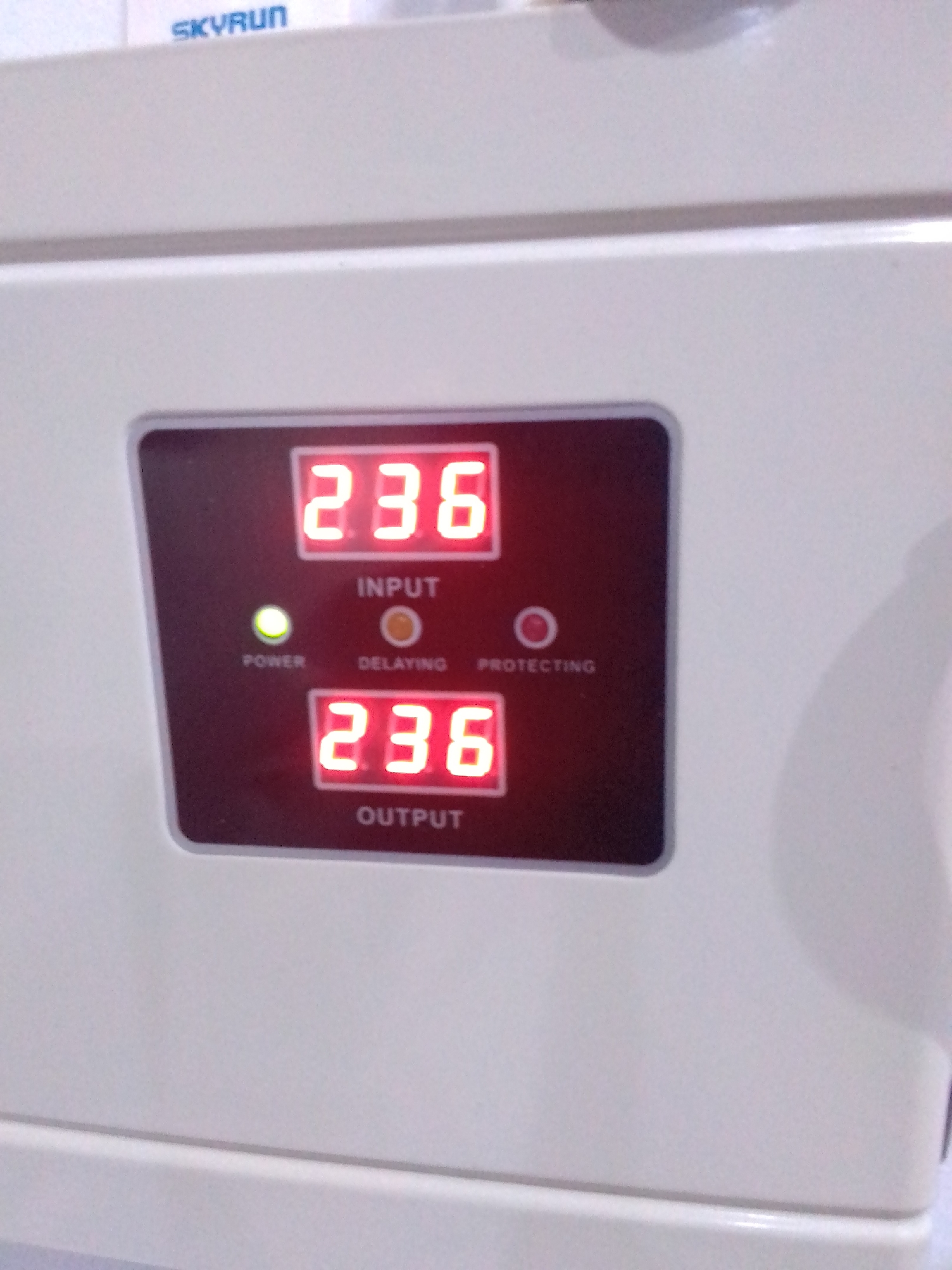 This media file is a close-up, eye-level shot of a white electronic device with a digital display. The device has a dark, rectangular panel on its front that houses two red LED displays, indicator lights, and labels. The top LED display shows "236" and is labeled "INPUT." Below this, there are three small circles, one green labeled "POWER," one orange labeled "DELAYING," and one red, unlabelled, positioned between "DELAYING" and "PROTECTING." The bottom LED display also shows "236" and is labeled "OUTPUT." The overall impression is that of a voltage regulator or a similar power conditioning device. The lighting appears to be artificial, with a slight glare on the device's surface. The background is out of focus but appears to be a light-colored surface, possibly another piece of equipment. Visible text on the device includes "SKYRUN" in blue lettering at the top of the frame, along with "INPUT," "POWER," "DELAYING," "PROTECTING," and "OUTPUT" in white lettering on the dark panel. The device appears to be in operation, indicated by the illuminated "POWER" light and the numerical displays.